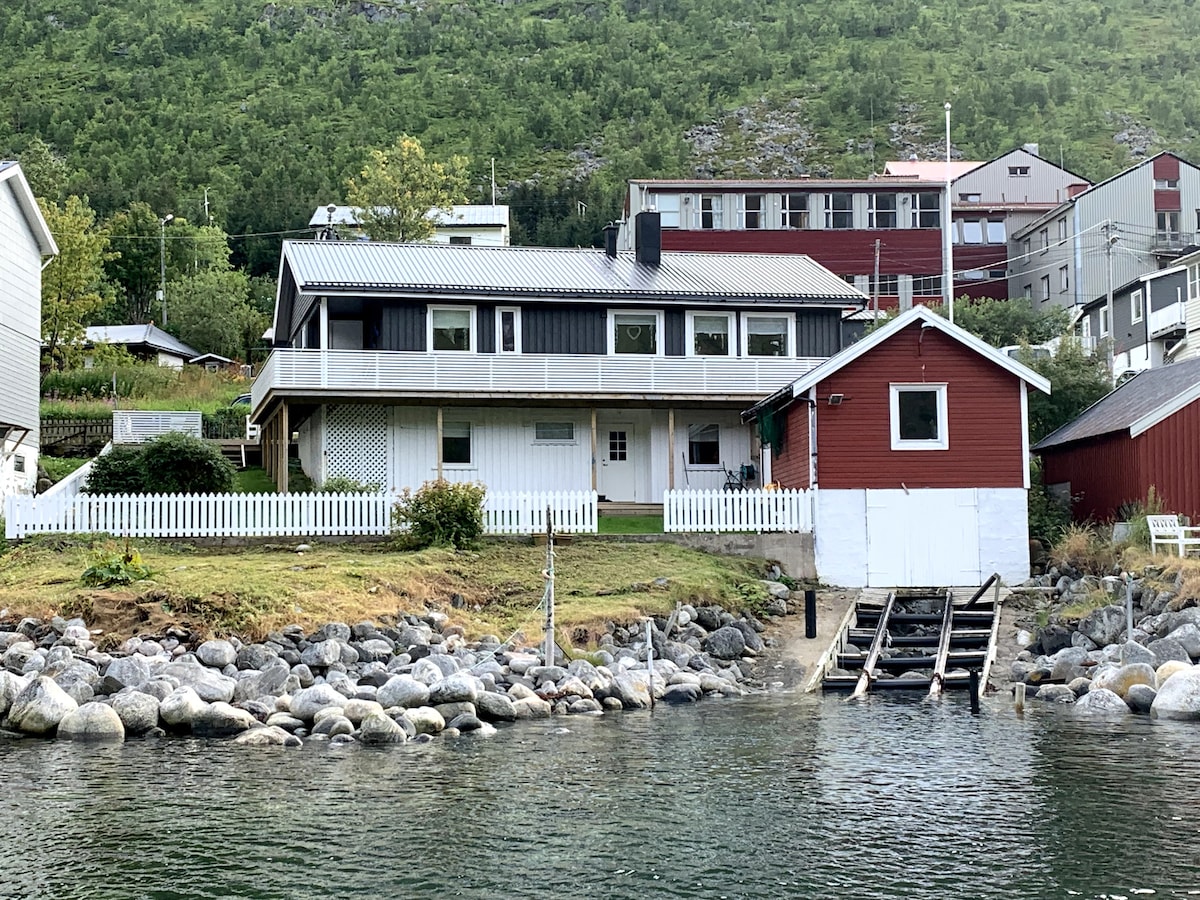 A scenic waterfront property is depicted, featuring a two-story house with a wooden balcony and a charming red building nearby. The landscape includes rocky shorelines leading to the calm water. Green hills rise in the background, enhancing the natural setting.