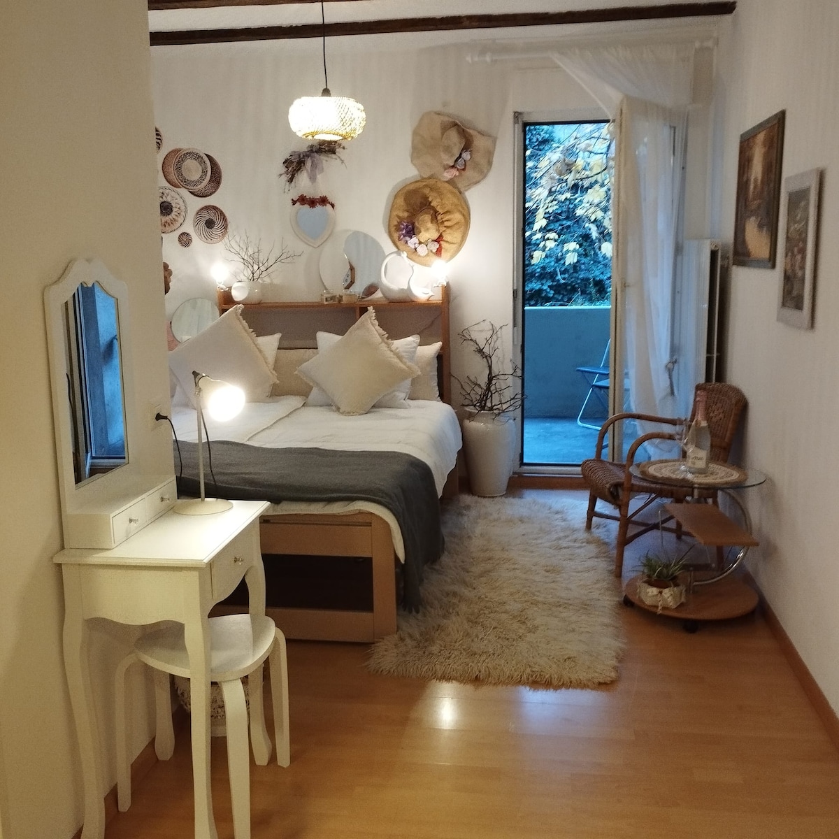 A cozy bedroom features a spacious bed adorned with decorative pillows. Natural light filters through a large glass door leading to a small outdoor area. A white desk with a lamp, a round table, and a rattan chair are positioned for convenience. Wall decorations and soft textures add warmth.