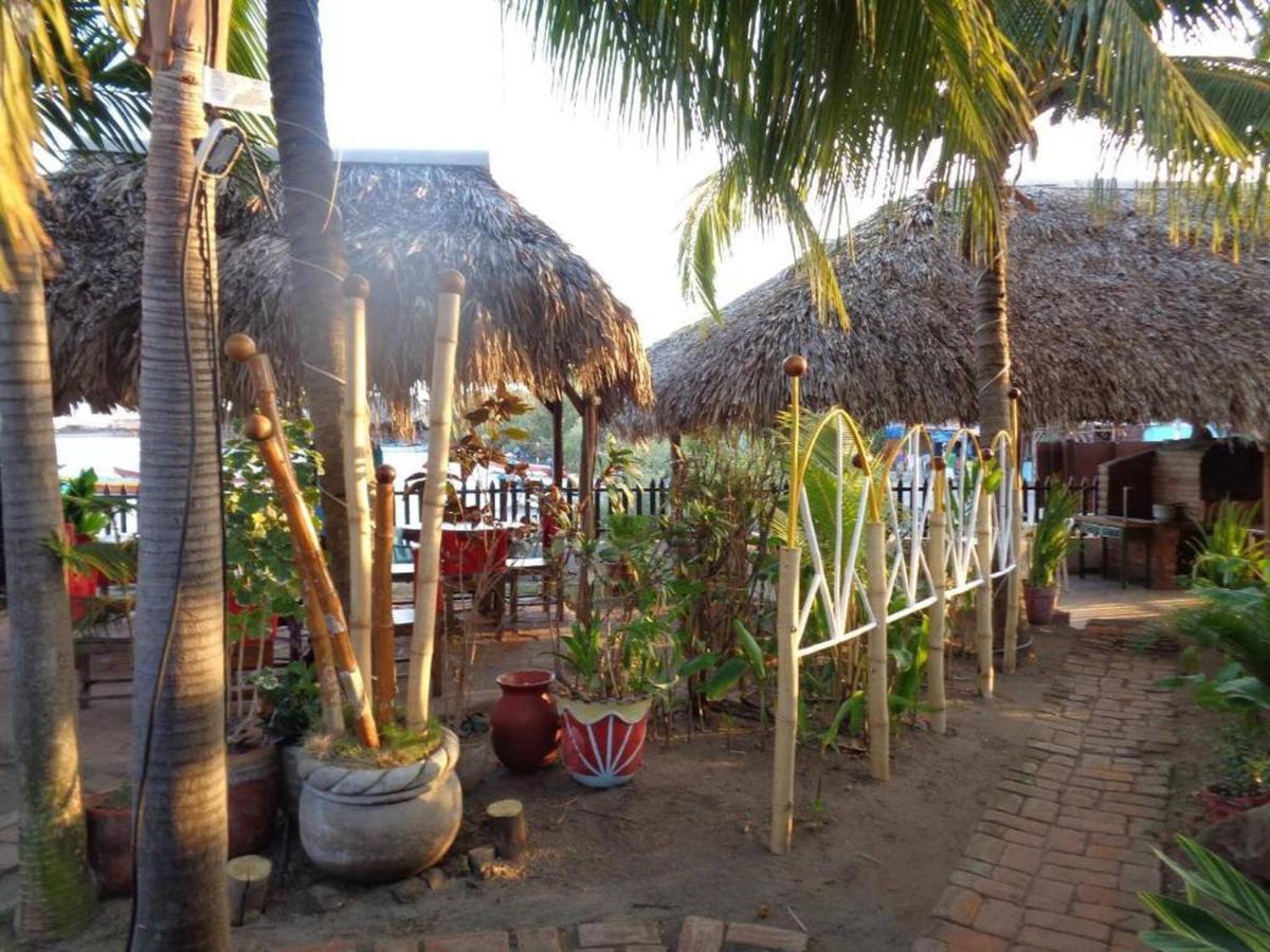 A tropical garden area is shown, featuring thatched-roof huts surrounded by palm trees and lush greenery. Pathways made of brick connect various seating areas, adorned with potted plants and decorative pottery, inviting guests to relax in a natural atmosphere.