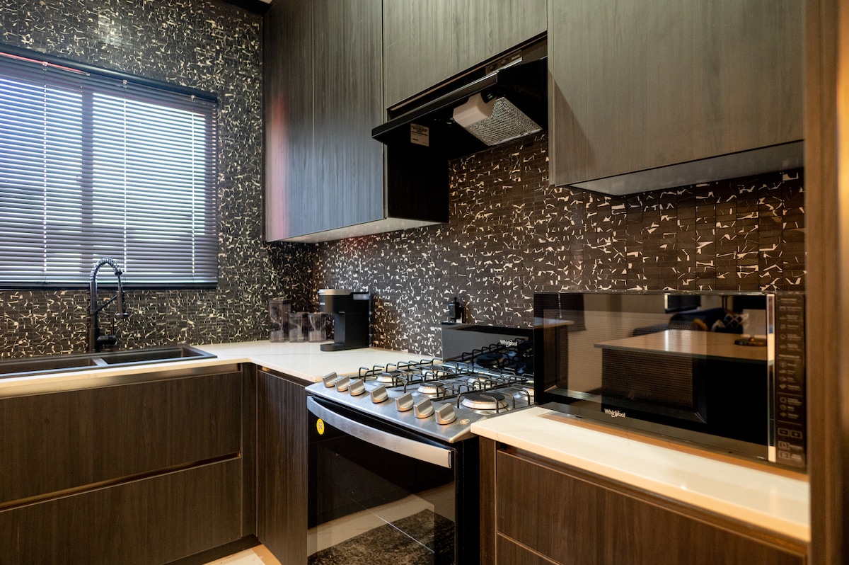 A modern kitchen features sleek dark cabinetry and a stylish mosaic backsplash. Stainless steel appliances, including a large gas stove and microwave, are present. Natural light filters in through a window, providing a bright atmosphere for cooking and meal preparation.