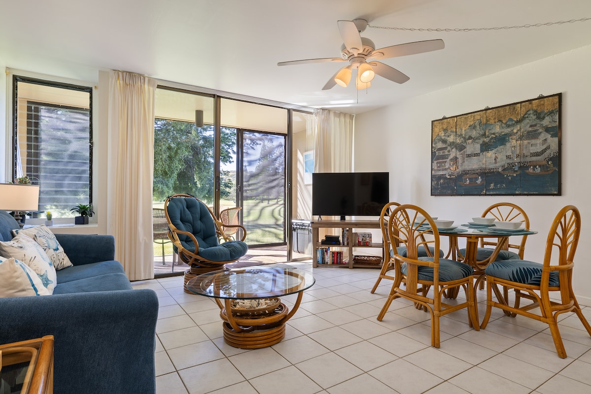 An open living area features a comfortable blue sofa, wicker furniture, and a glass coffee table. A dining table for four is positioned near large sliding glass doors that lead to the lanai, offering views of the lush surroundings. Bright natural light fills the space.