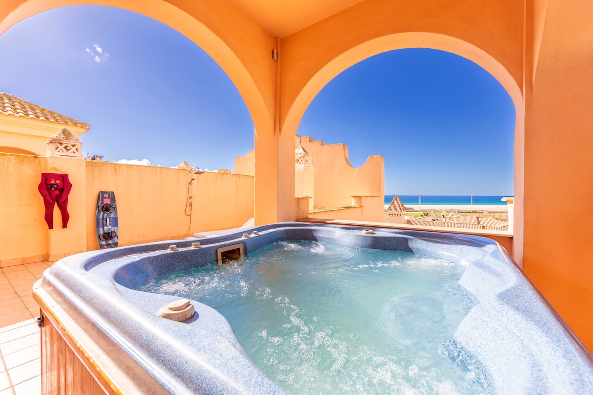A private jacuzzi is situated on a terrace with an ocean view. The spa area features smooth, tiled surroundings and is framed by arched openings that allow natural light and outdoor scenery to be enjoyed. The coastline and blue skies are visible in the background.