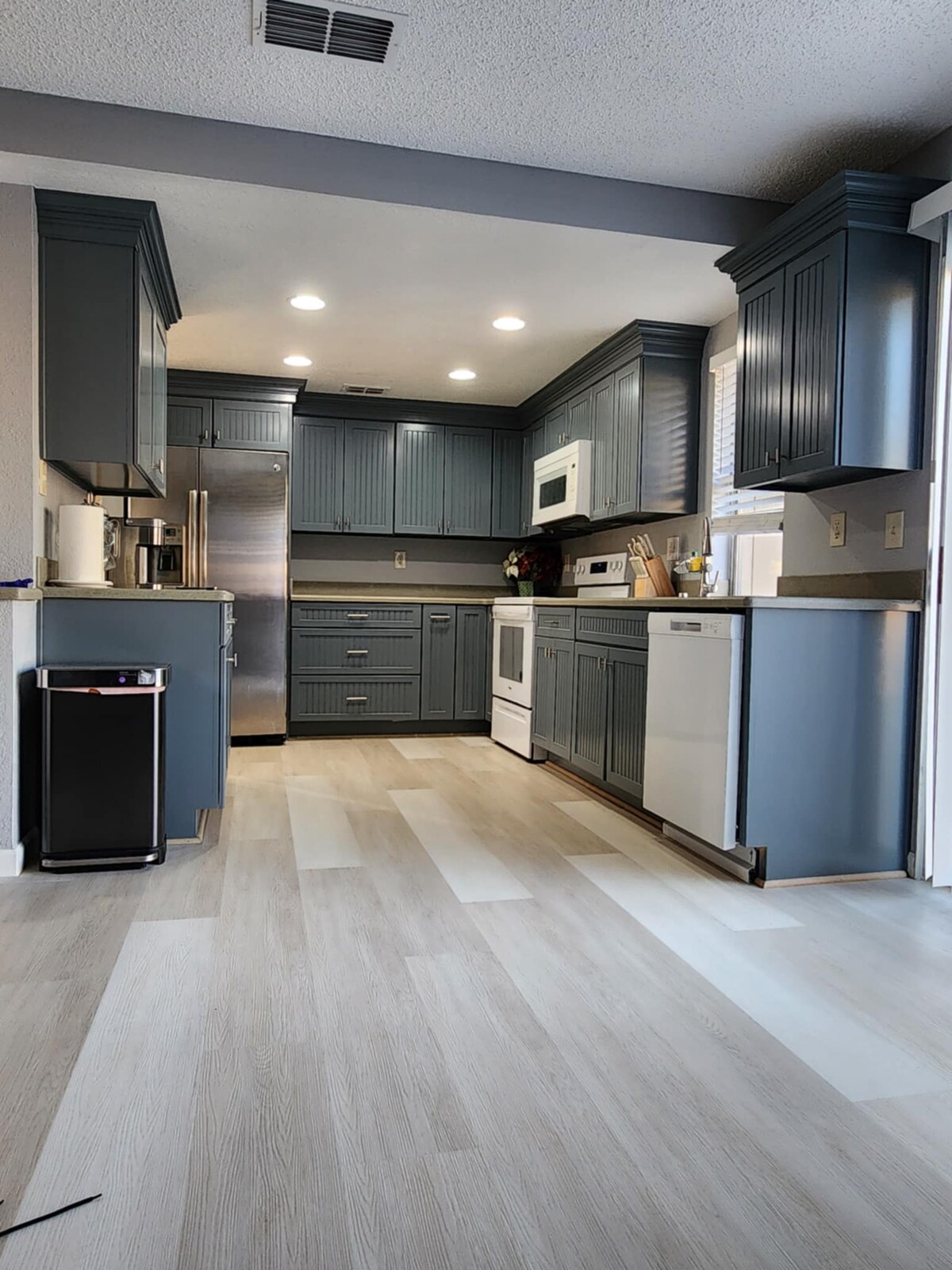 A modern kitchen features navy blue cabinetry and stainless steel appliances. Natural light illuminates the space from a nearby window. A white stove and microwave create a cohesive look, while light-colored flooring adds brightness. A small trash can stands near the entrance for convenience.