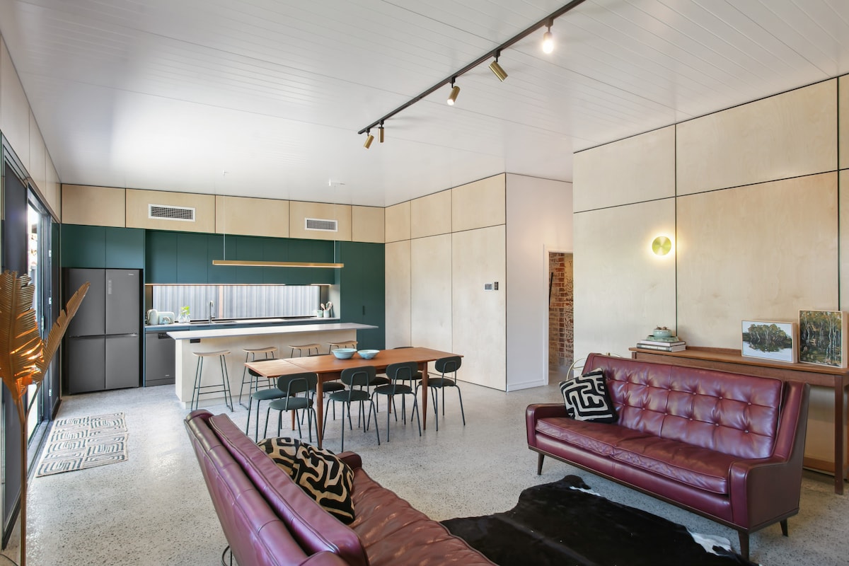 An open-plan living area showcases a modern kitchen with green cabinetry and stainless steel appliances. A wooden dining table is surrounded by dark chairs. Two burgundy sofas offer seating, while the concrete floor is complemented by subtle decor elements and wall features of exposed brick.