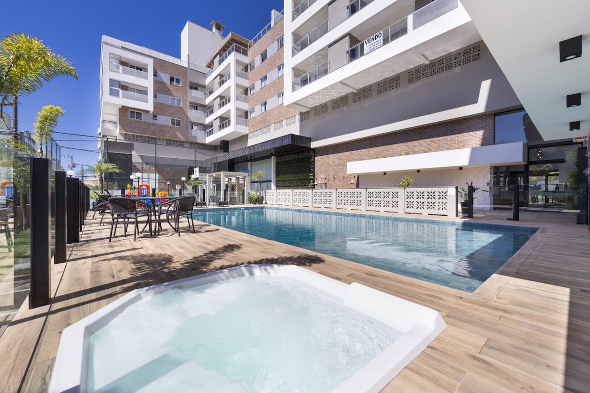 An outdoor area features a swimming pool surrounded by sun loungers and a dining set with dark chairs and a table. A hot tub is positioned in the foreground. The clean lines of the modern building and palm trees create an inviting atmosphere.
