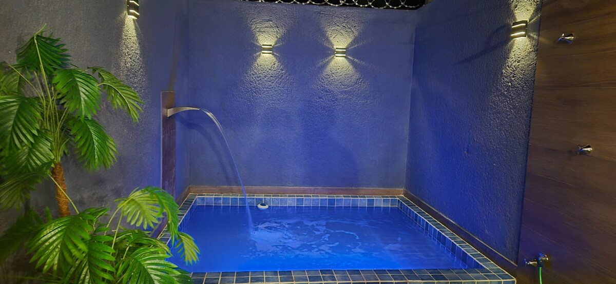 A small private pool is set against a textured blue wall, softly illuminated by wall-mounted lights. A gentle water feature adds to the serene ambiance, complemented by a lush potted plant positioned at the edge.