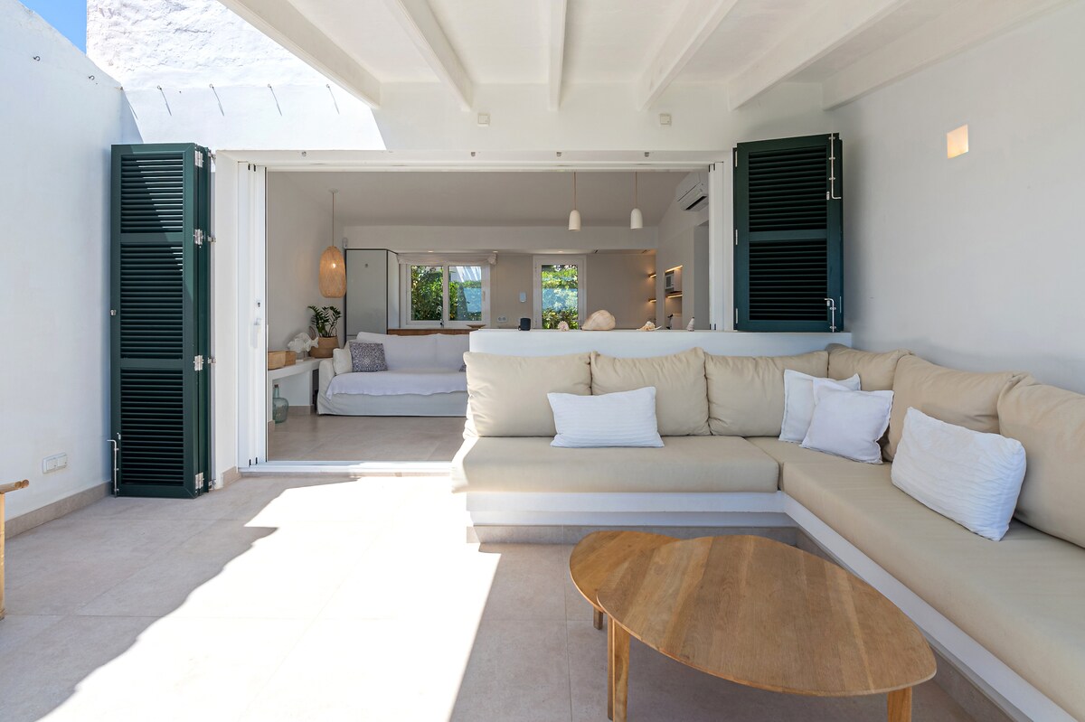 A spacious living area features a light-colored sectional sofa with multiple decorative pillows and a wooden coffee table. Large glass doors lead to the outdoor terrace, allowing natural light to fill the room. Minimalist decor creates a serene environment.