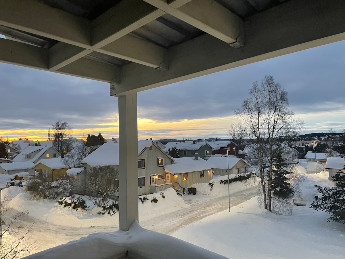 A view from the balcony showcases a snowy landscape, with houses nestled within the white surroundings. The sunset casts soft hues across the sky, highlighting the tranquil atmosphere. Snow-covered rooftops and trees frame the scene, suggesting a peaceful neighborhood.