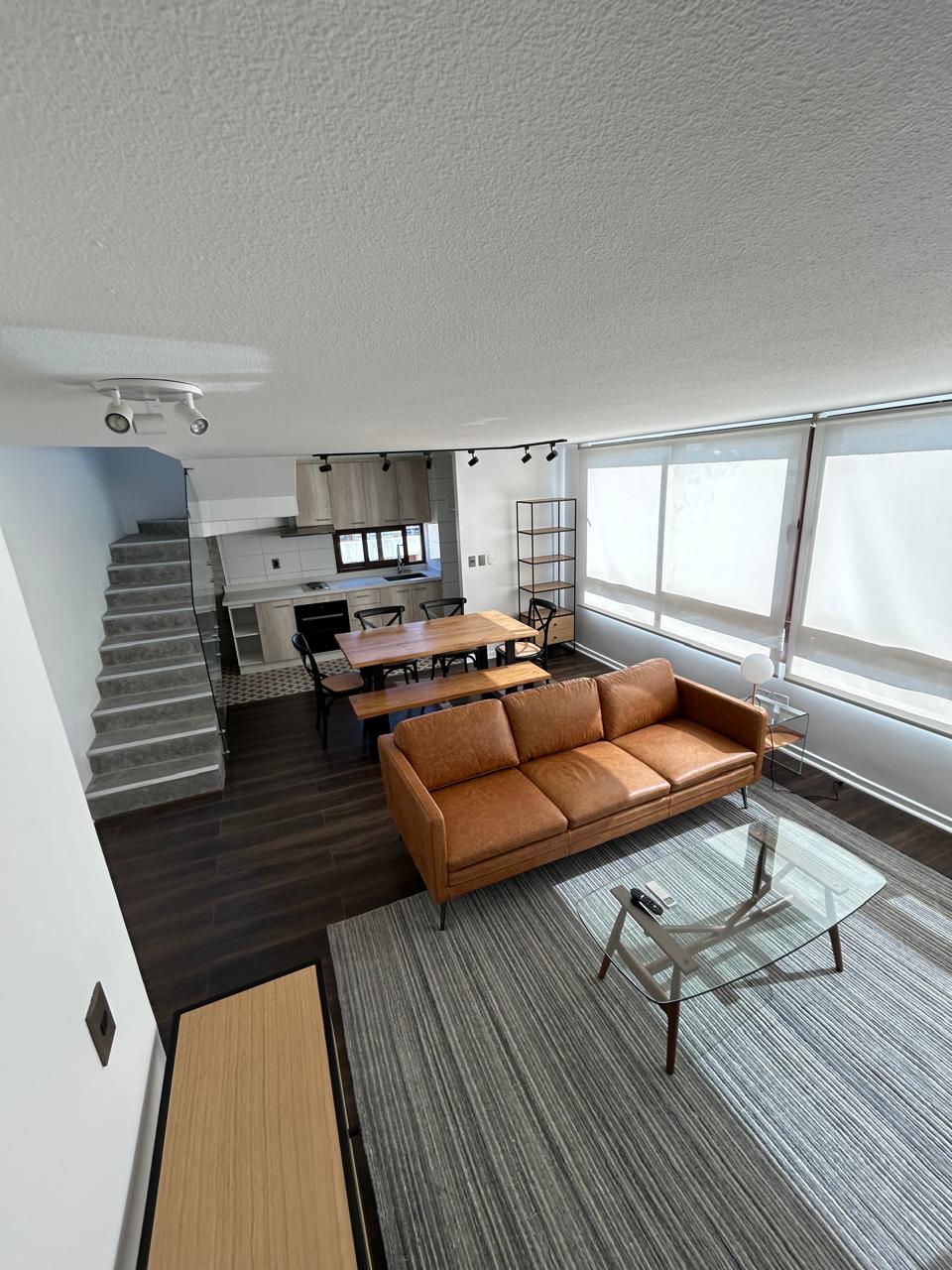 An open living area displays a modern sofa and a glass coffee table, complemented by a wooden dining table that seats six. Natural light filters through large windows with roller shades. The sleek kitchen area is visible in the background, with a staircase leading to the upper level.