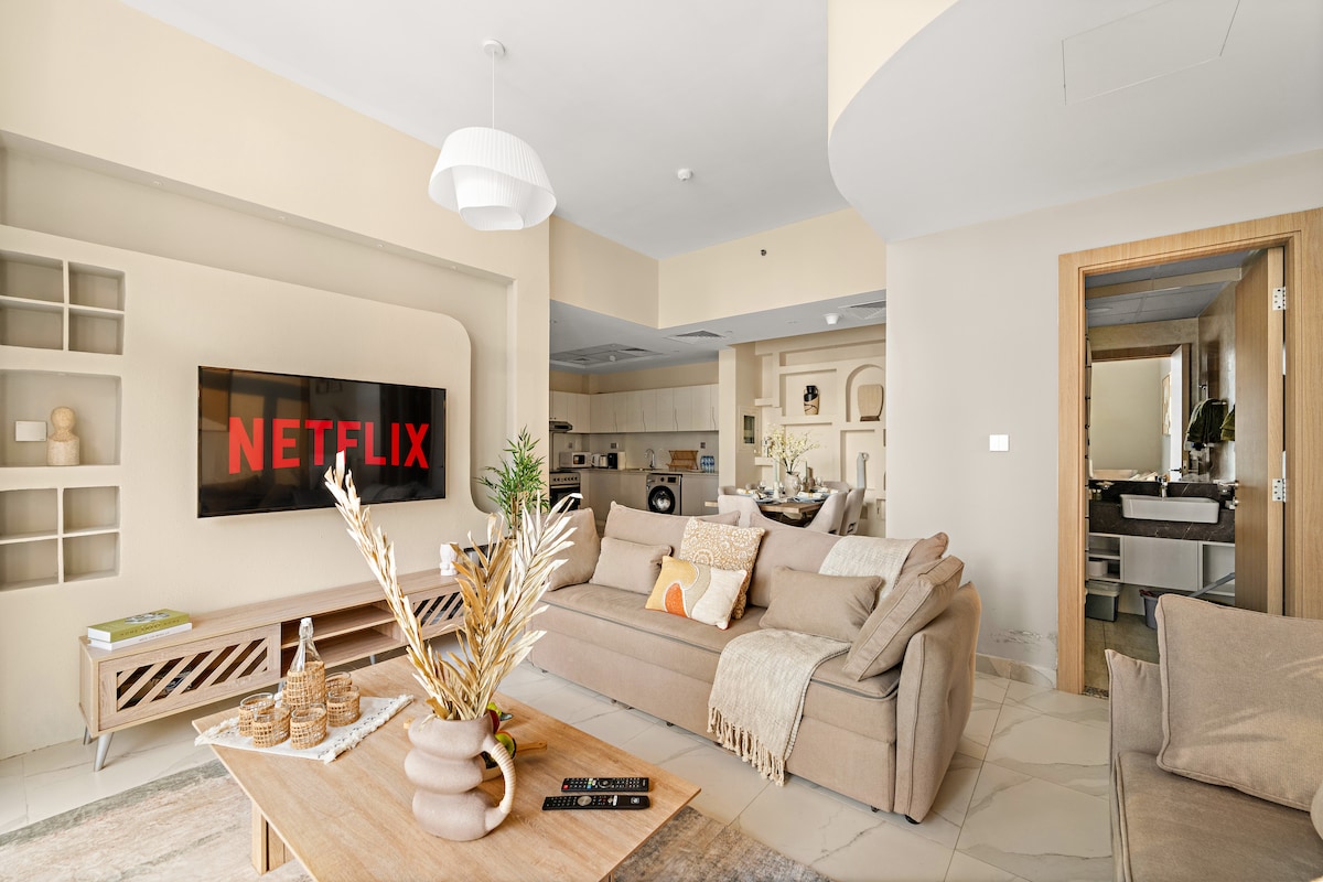 A modern living room is featured with a spacious sofa adorned with textured cushions. A wall-mounted television displays the Netflix logo. A wooden coffee table holds decorative elements, while the open layout connects to the kitchen and dining area in the background.