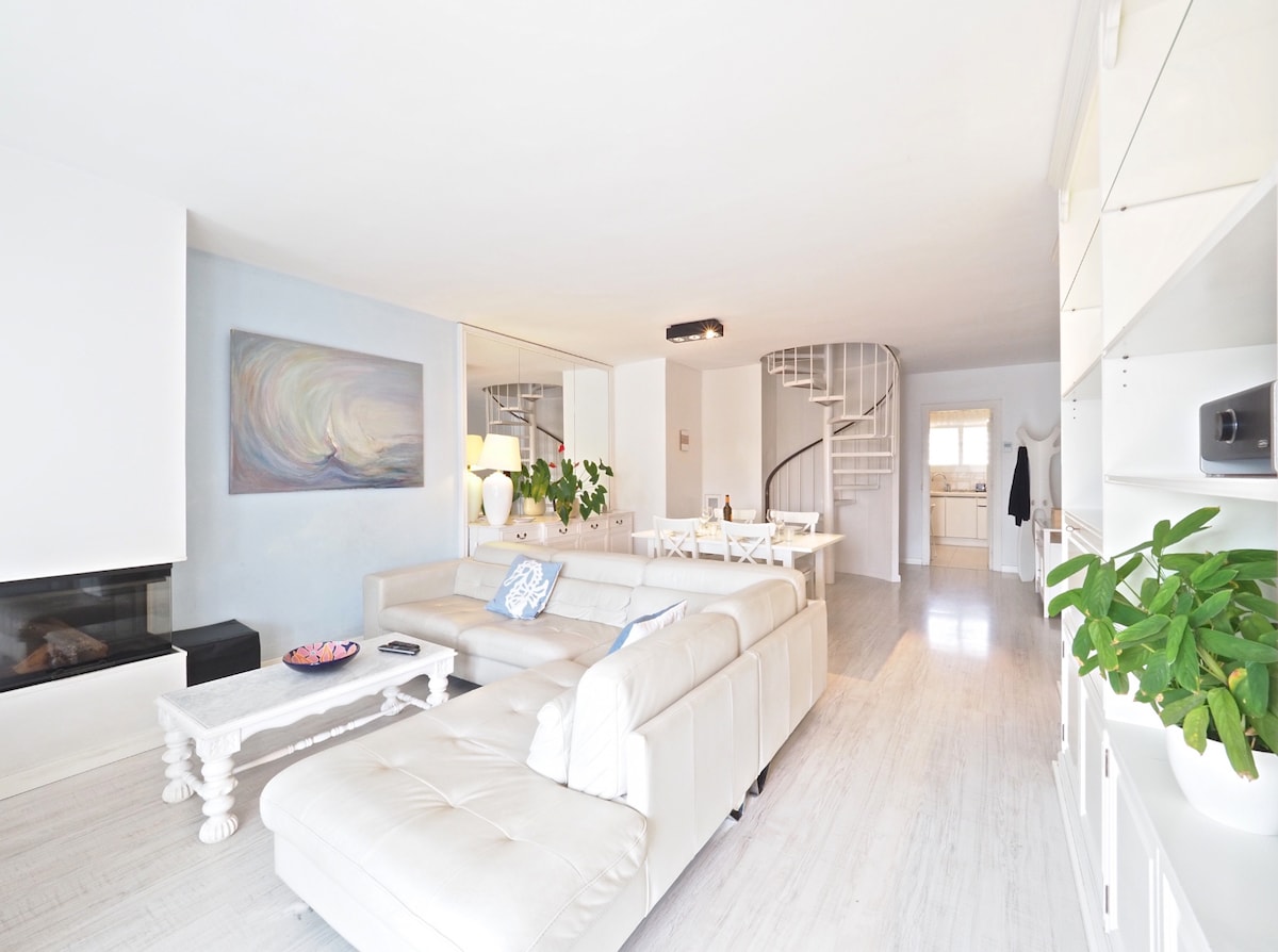 A spacious living area features a light-colored sofa with a chaise longue, complemented by a decorative coffee table. A vibrant piece of artwork adorns the wall, while plants enhance the freshness. A dining table for eight is visible, along with a staircase leading to the upper level.