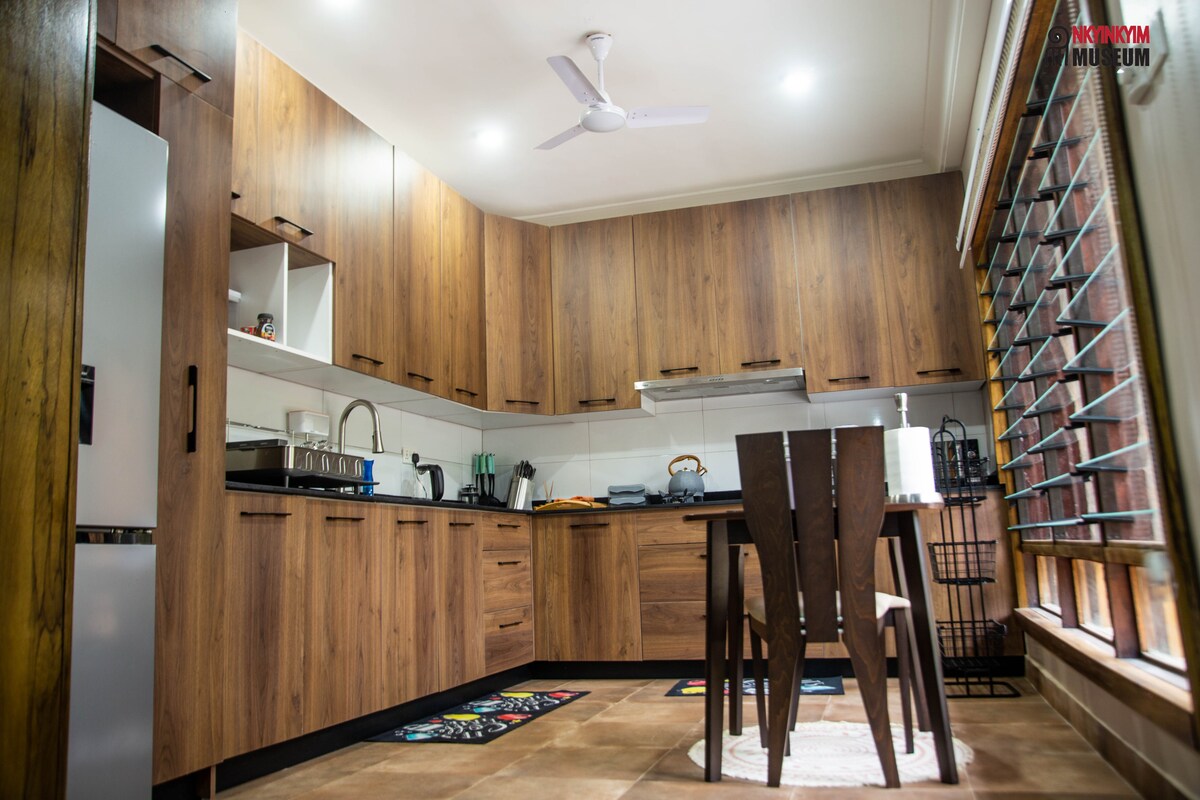 A modern kitchen features warm wooden cabinetry and a white countertop. A dining table set for four is positioned nearby. Large windows provide natural light, and a ceiling fan enhances air circulation. Appliances are neatly arranged, contributing to a functional cooking space.