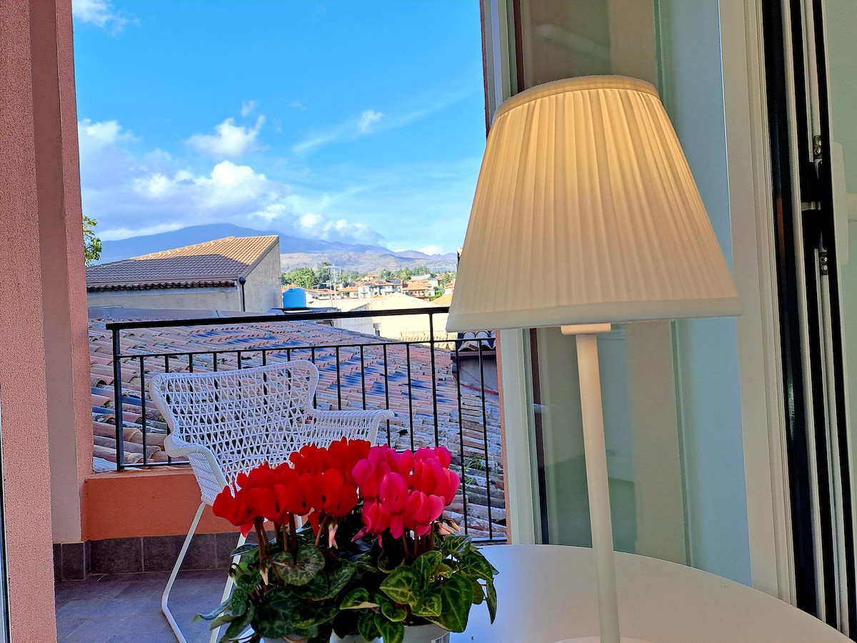 A balcony view is presented, featuring colorful flowers in a planter on a table. A white lamp stands beside the flowers. The skyline reveals rooftops and distant mountains under a partly cloudy blue sky.