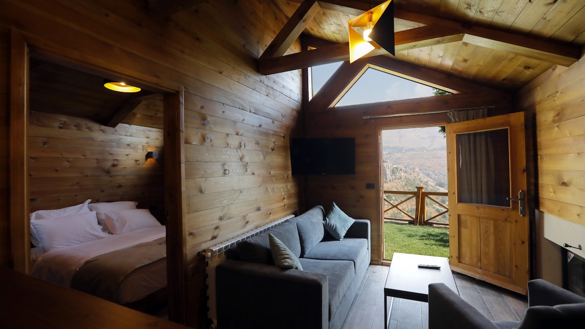 The image showcases a wooden lodge interior, featuring a cozy seating area with a sofa facing a coffee table. A spacious bed is visible in the adjacent room. Large windows allow natural light to flow in, framing views of the surrounding landscape.