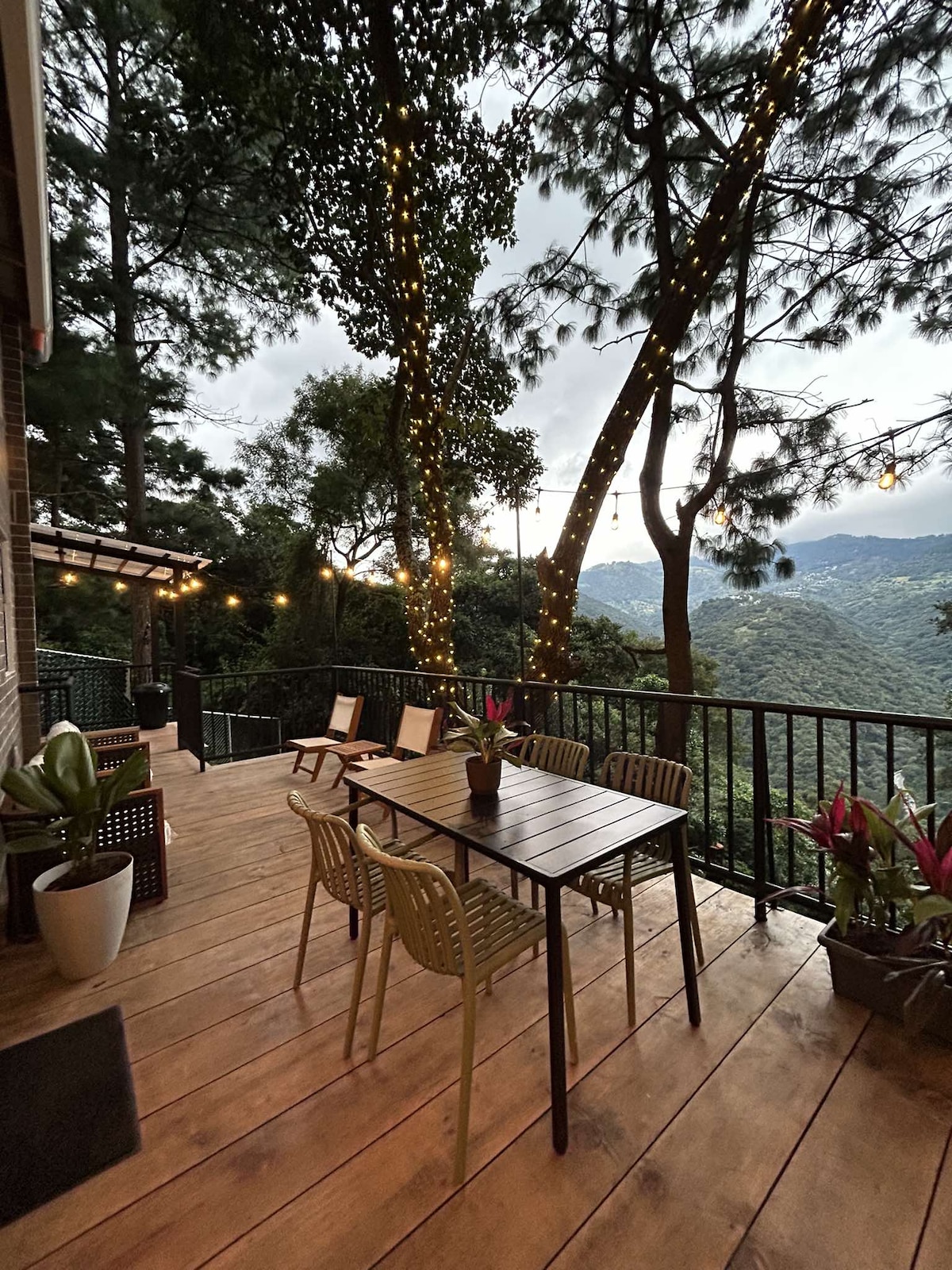 An outdoor terrace features a wooden deck with a dining table and four chairs. Surrounding greenery adds a natural touch, while string lights create a soft glow. The mountainous landscape provides a scenic backdrop, enhancing the serene atmosphere of the space.