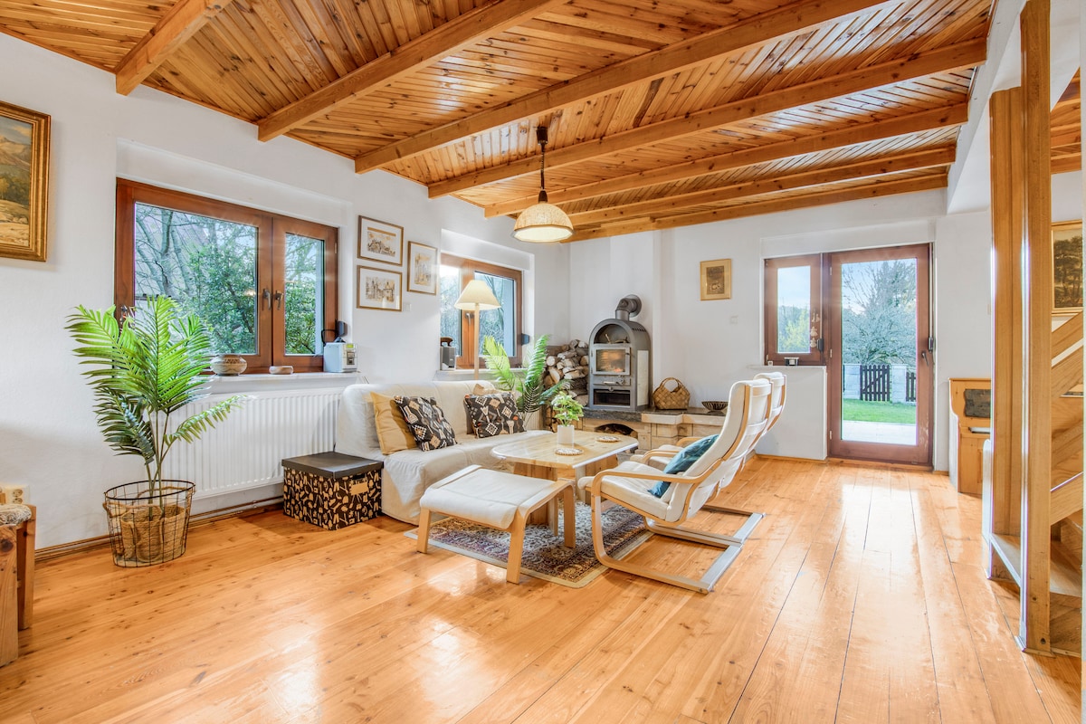 A living area features wooden beams and a warm wooden floor. A cozy seating arrangement includes a pair of chairs and a low table. Natural light enters through large windows, illuminating a decorative plant nearby. A fireplace adds a touch of comfort to the space.