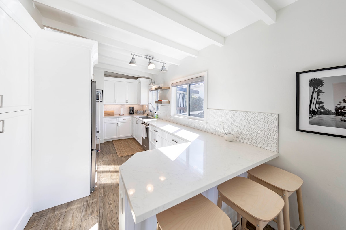 A bright kitchen features white cabinetry and modern stainless steel appliances. A spacious island with a white countertop provides seating for three on light wooden stools. Large windows allow light to fill the space, highlighting sleek lines and a clean design.