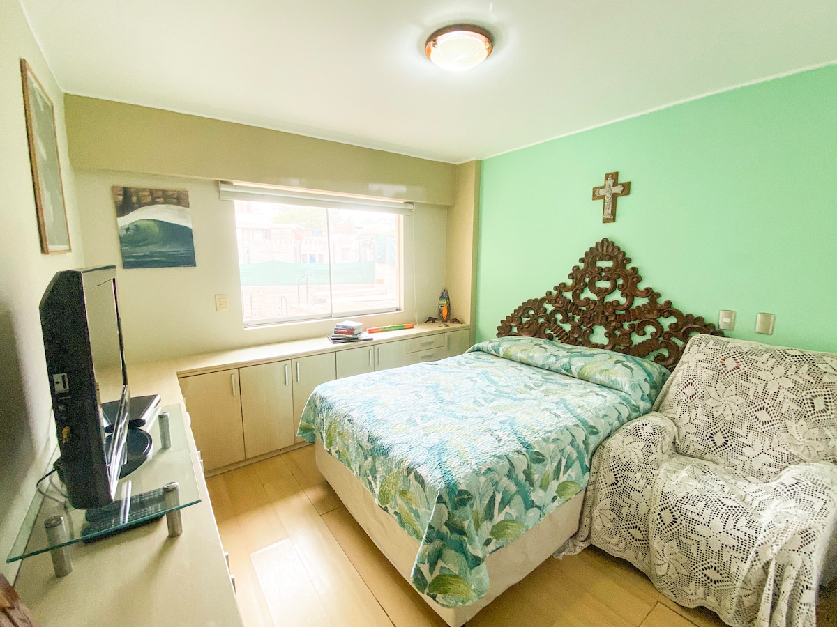 A thoughtfully arranged bedroom features a queen-sized bed with a vibrant blue and green bedspread. A small TV is positioned on a light-colored media unit across from the bed. Natural light enters through a window, complemented by a decorative cross and a cozy armchair in the corner.