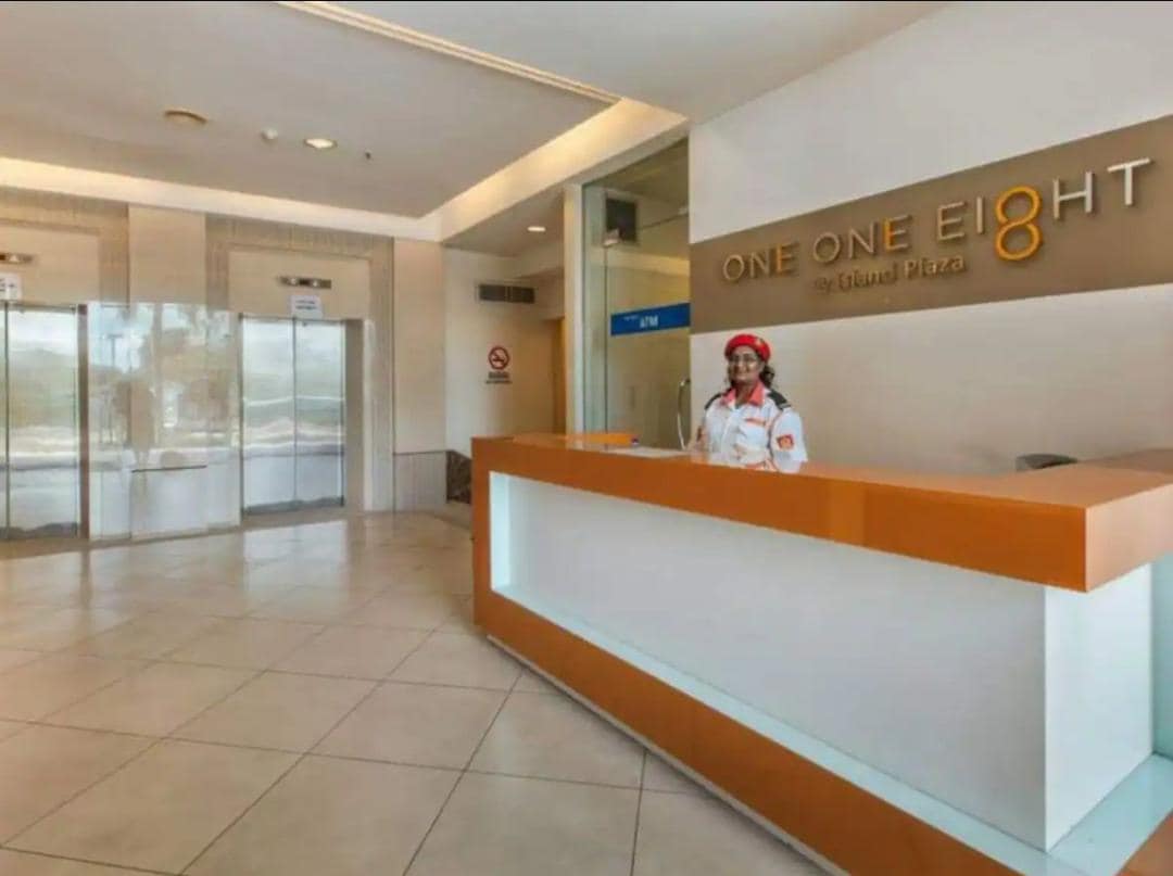 The reception area is designed with a modern aesthetic, featuring a sleek front desk and large glass doors. Natural light fills the space, highlighting the tiled floor and welcoming atmosphere. A friendly staff member is present, ready to assist guests.