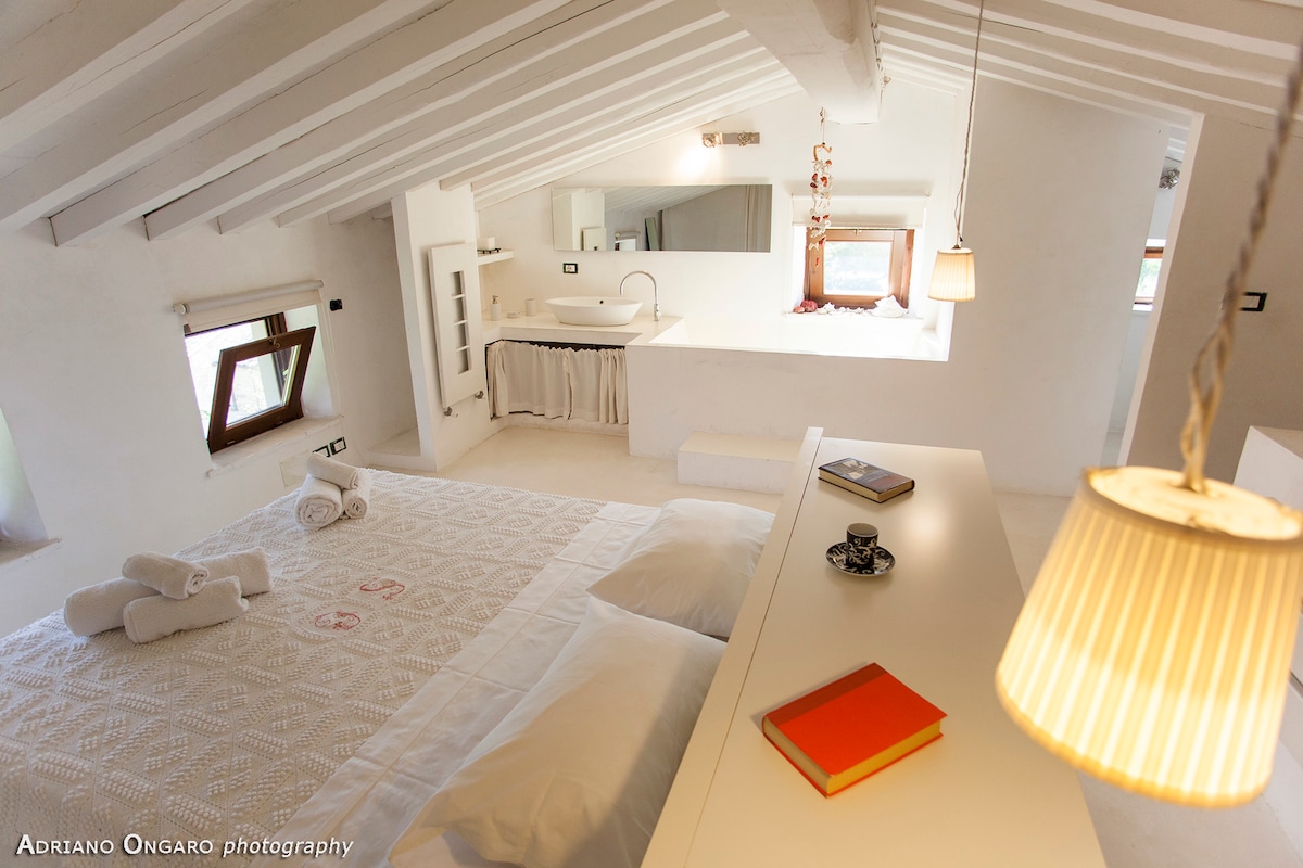 A bright, airy room features a spacious bed covered with a textured blanket and neatly arranged towels. A small table holds a decorative cup and a book. Natural light enters through windowed walls, and subtle ceiling beams enhance the open feel of the space.
