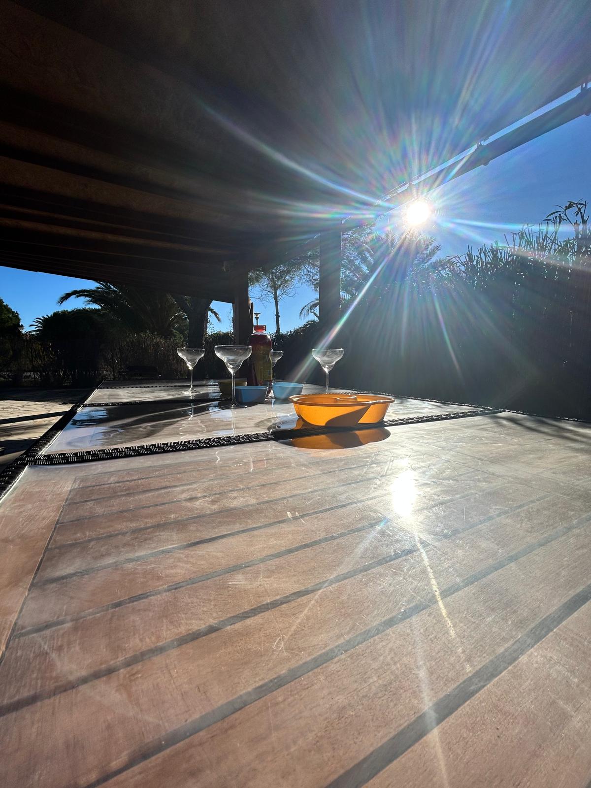 A spacious outdoor terrace is featured, bathed in sunlight. A glass of wine and a small bowl are placed on a table, reflecting the bright rays. Lush greenery can be seen in the background, enhancing the serene atmosphere of the space.