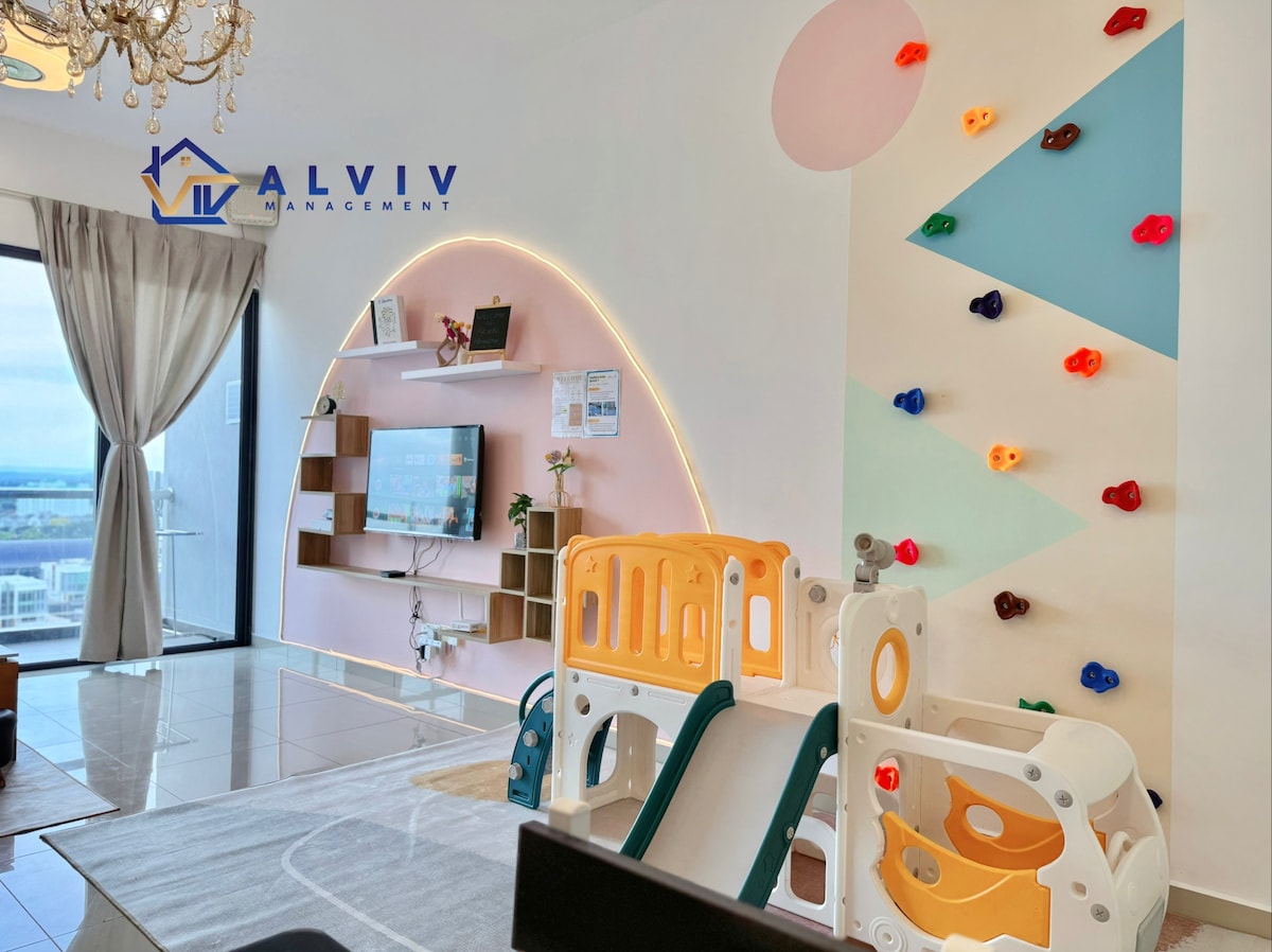 A bright living area features a playful children's climbing wall adorned with colorful shapes. An entertainment unit is mounted against a curved wall with soft lighting, creating an inviting space. A multi-colored play structure is positioned nearby, designed for young children in a family-friendly environment.