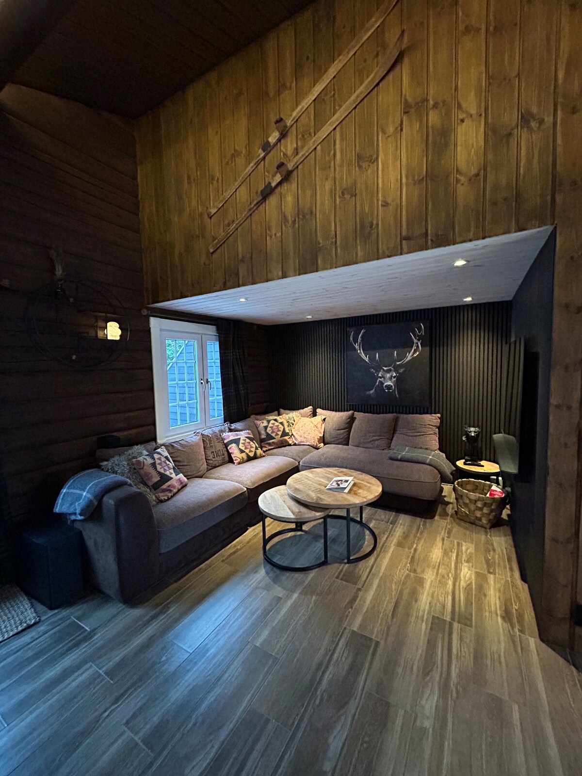 A corner seating area features a spacious sectional sofa adorned with decorative pillows. A round coffee table is positioned in front, and warm wood paneling complements the overall design. Natural light enters through the window, creating a cozy ambiance.