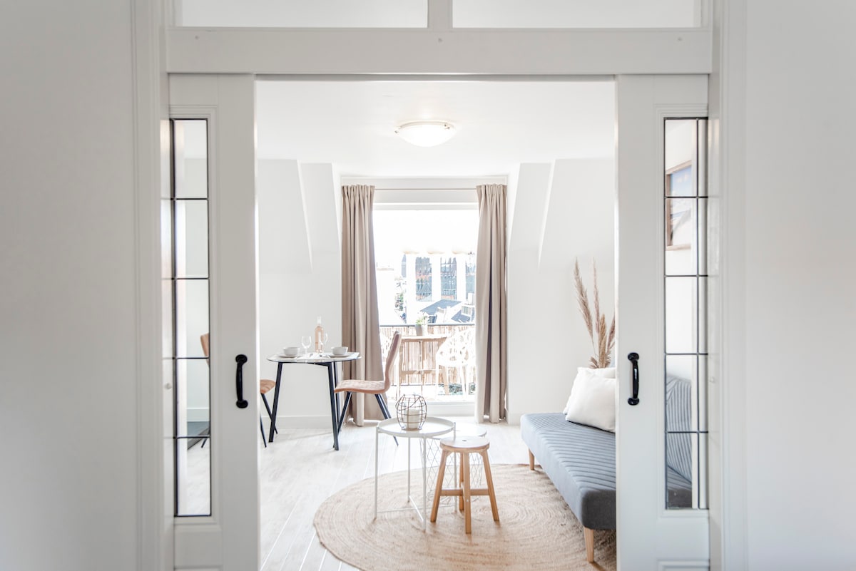 A bright living area is viewed through glass paneled doors. It features a cozy grey couch positioned across from a round table set for two. Natural light floods the space, highlighting light-colored flooring and neutral-toned curtains. A balcony is visible in the background.