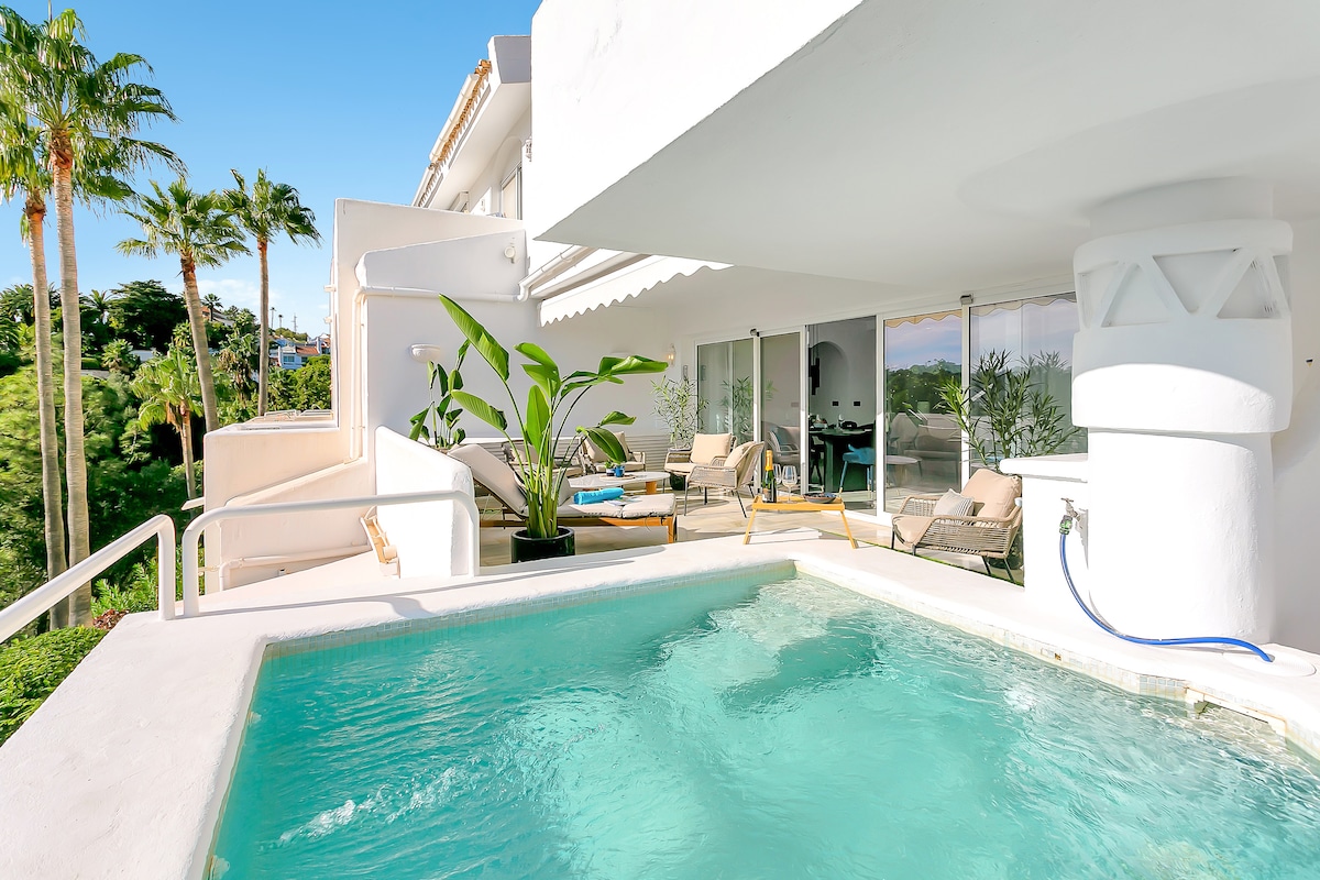 A private terrace showcases a splash pool surrounded by lounge chairs and tropical plants. The bright and spacious area is illuminated by sunlight, offering views of the lush greenery beyond. Sliding glass doors provide easy access to the indoor living space.