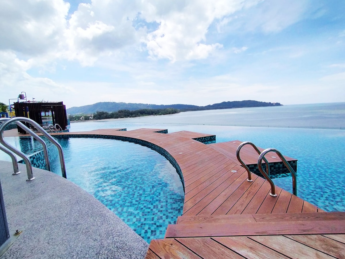 A swimming pool extends towards the horizon, featuring a curved design with wooden decking. The serene water reflects the sky, while hills are visible in the distance, creating a sense of openness and tranquility.