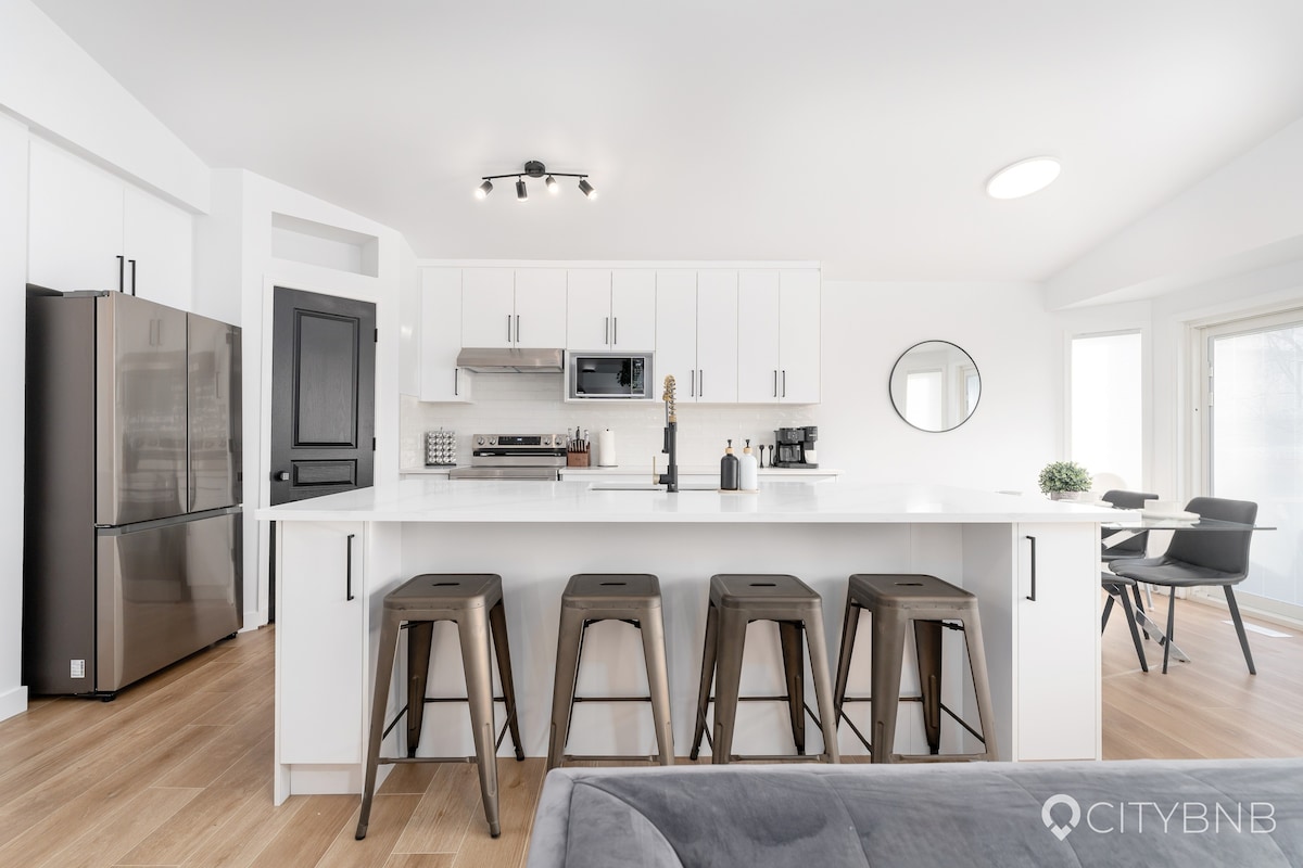 A modern kitchen features white cabinetry and a central island with seating for four. Stainless steel appliances, including a refrigerator, are visible. Natural light streams in through large windows, illuminating the open space and highlighting a round mirror on the wall.