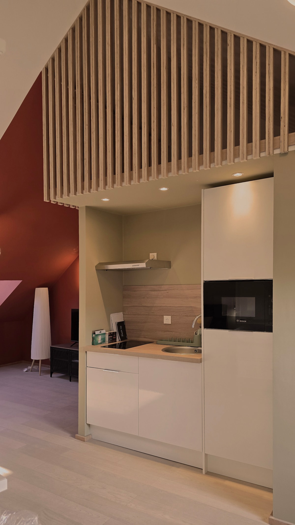 A modern kitchen area is presented with sleek cabinetry and integrated appliances. Neutral tones are complemented by wooden slat accents above, adding an element of texture. Soft lighting enhances the functional space, which includes a sink and stovetop, alongside ample counter space for meal preparation.