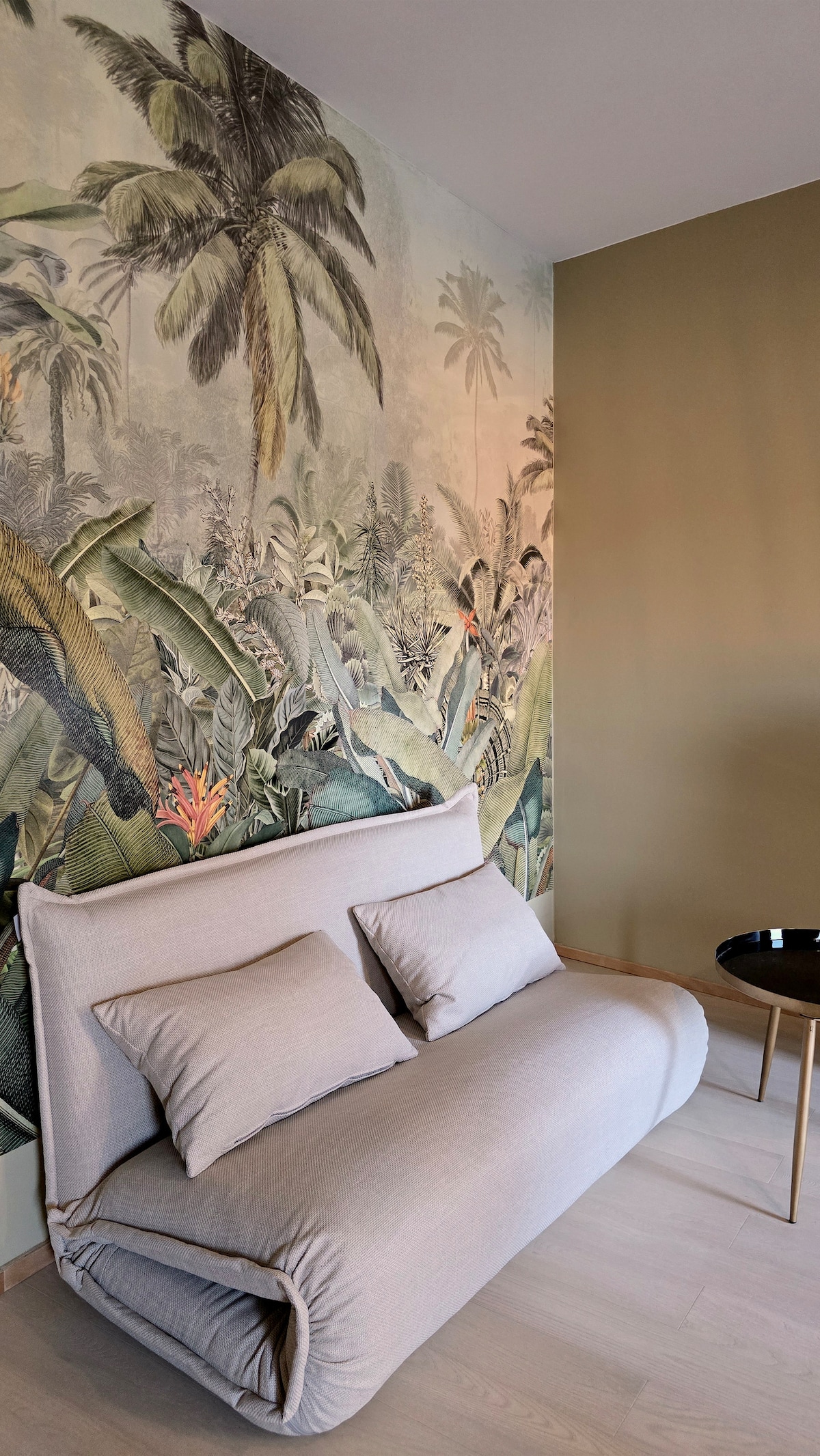 A relaxing seating area features a foldable sofa with two plush cushions. The wall behind showcases a vibrant tropical mural depicting lush greenery and palm trees. A small black side table sits nearby, complementing the earthy tones of the adjacent wall.