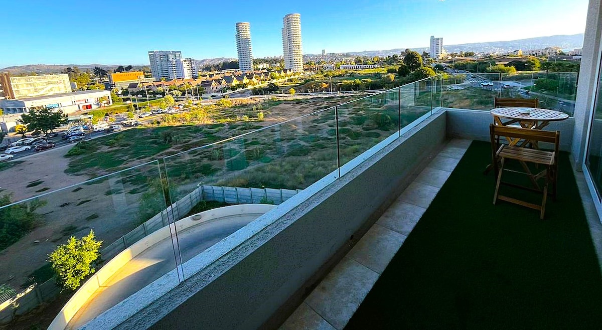 A spacious balcony features a small wooden table with two folding chairs, offering a view of the surrounding cityscape. Glass railings provide an unobstructed sightline, while a patch of green grass adds a touch of nature to the outdoor space.