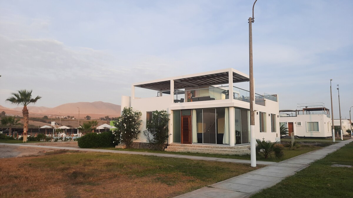 The modern two-story house features large glass windows and a spacious terrace on the rooftop. Surrounded by well-maintained greenery, the exterior presents a clean and inviting appearance. The mountainous backdrop enhances the tranquil setting of this coastal property.