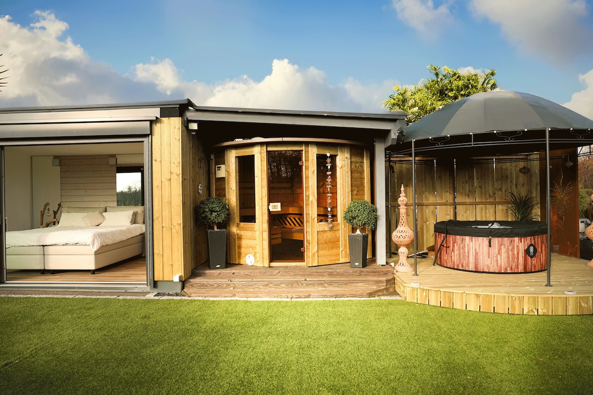 A wooden structure features a modern bedroom with large glass doors opening onto a wooden deck. Adjacent to the bedroom, a sauna is visible, and a round wooden hot tub is nestled under a gazebo, surrounded by manicured grass.