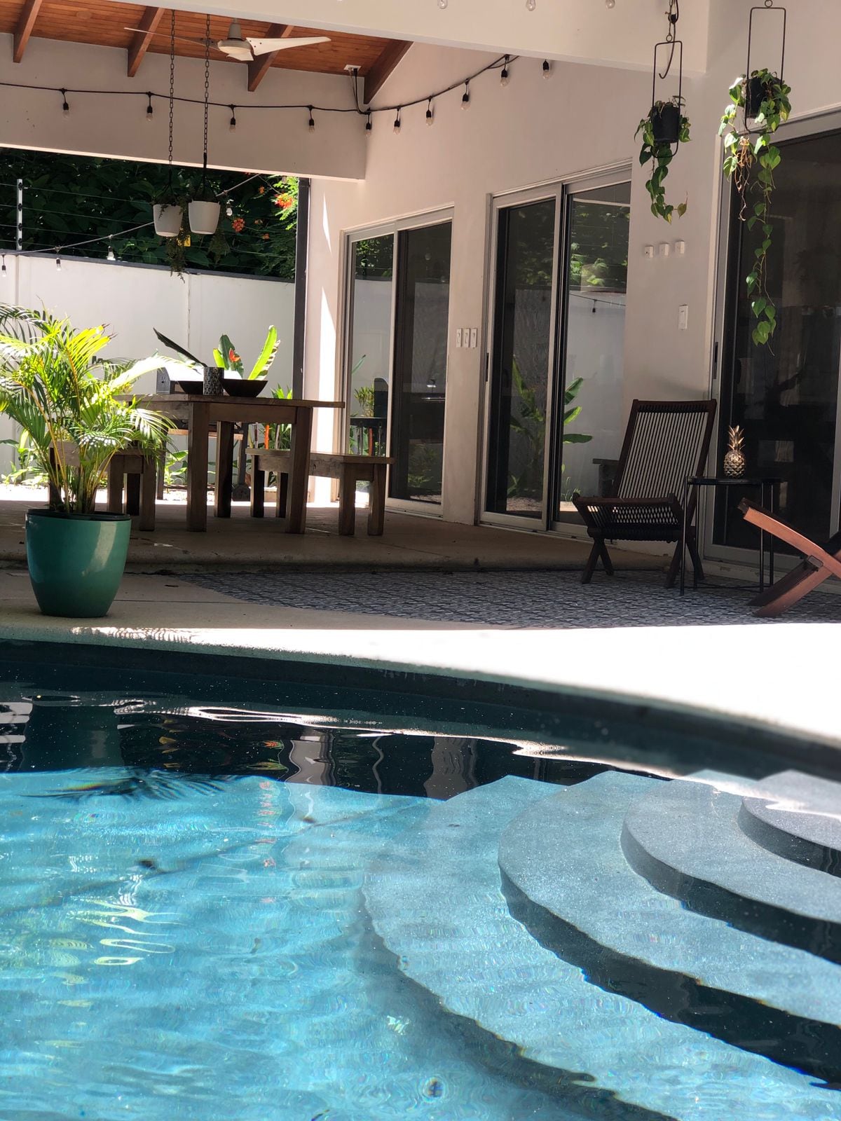 A clear view of a swimming pool with steps leading into the water is presented. The outdoor area features a dining table and chairs nearby, complemented by potted plants. Large glass doors allow natural light to fill the space, enhancing the connection with the landscaped yard.