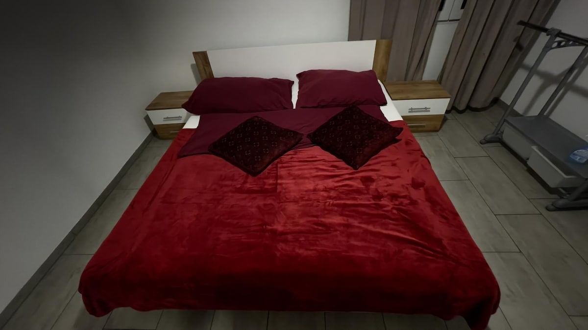 A double bed is dressed in a deep red blanket, complemented by maroon pillows. Nightstands are positioned on either side, and a folding rack is visible in the corner. Soft light filters in through curtains, enhancing the cozy feel of the space.