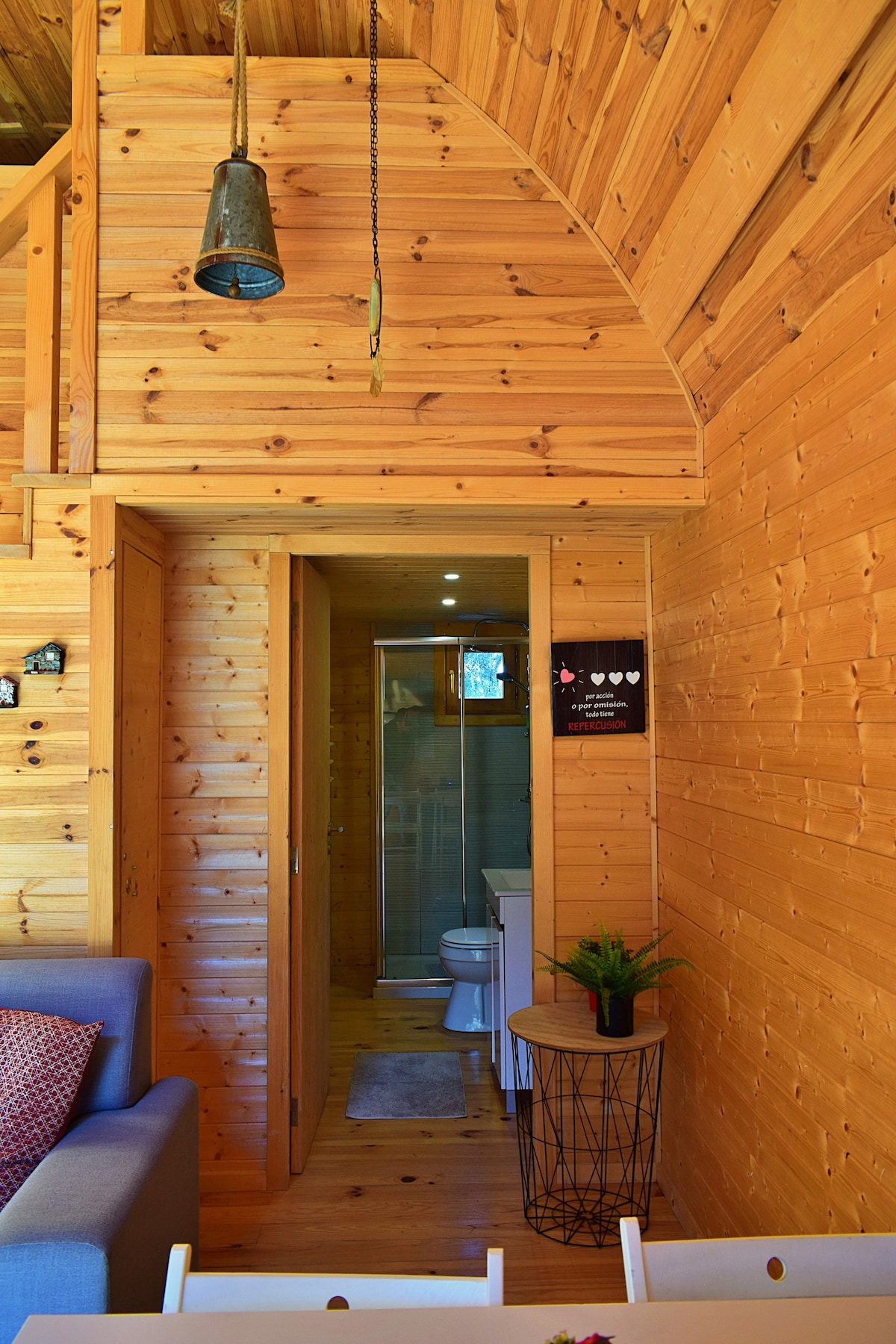The interior space features warm wooden paneling and an arched ceiling, creating a cozy atmosphere. A bathroom door is visible, leading to a private restroom. A comfortable sofa sits nearby, accompanied by a circular side table adorned with a small plant.