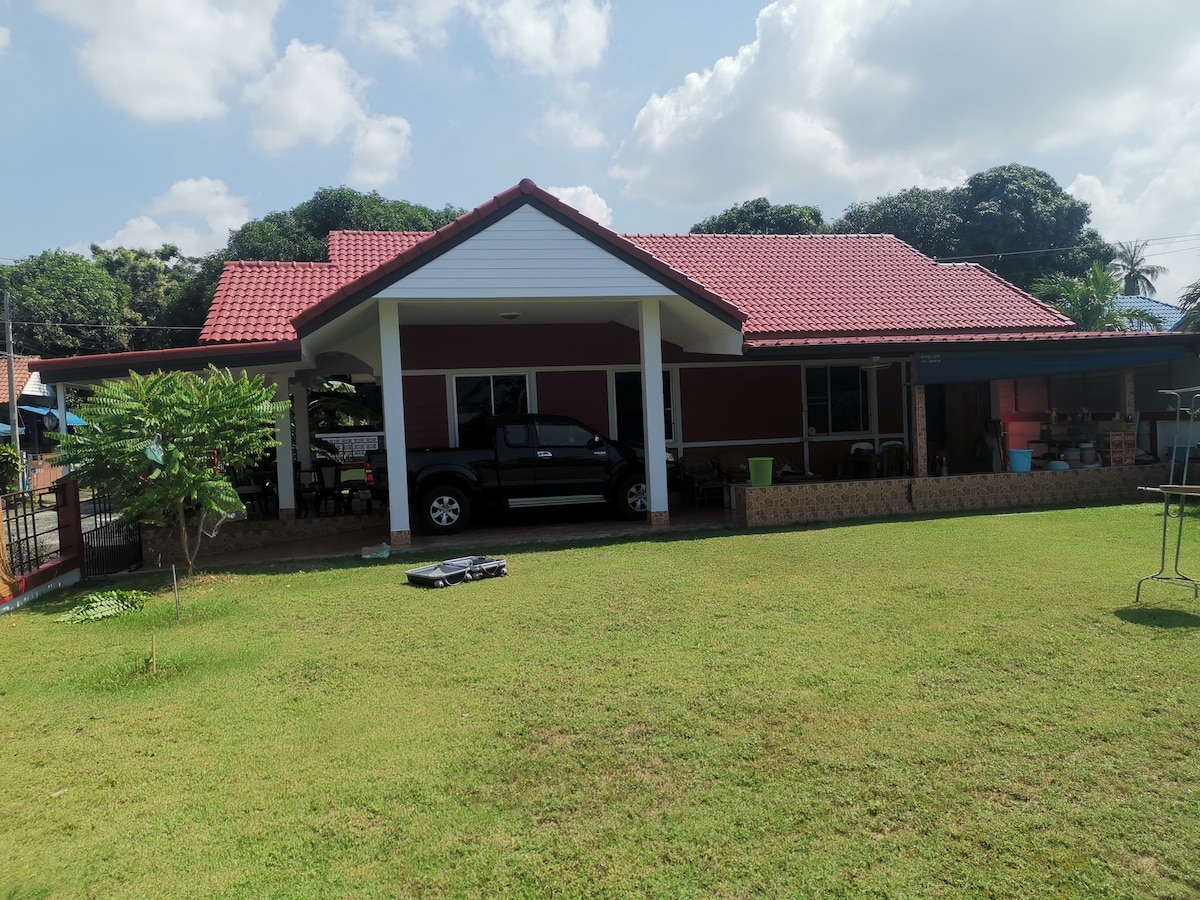 The single-story house features a red-tiled roof and a large front yard, with well-maintained grass. A black vehicle is parked under a covered area, while an outdoor seating space is visible beside the house. Lush trees and foliage surround the property, providing a serene environment.