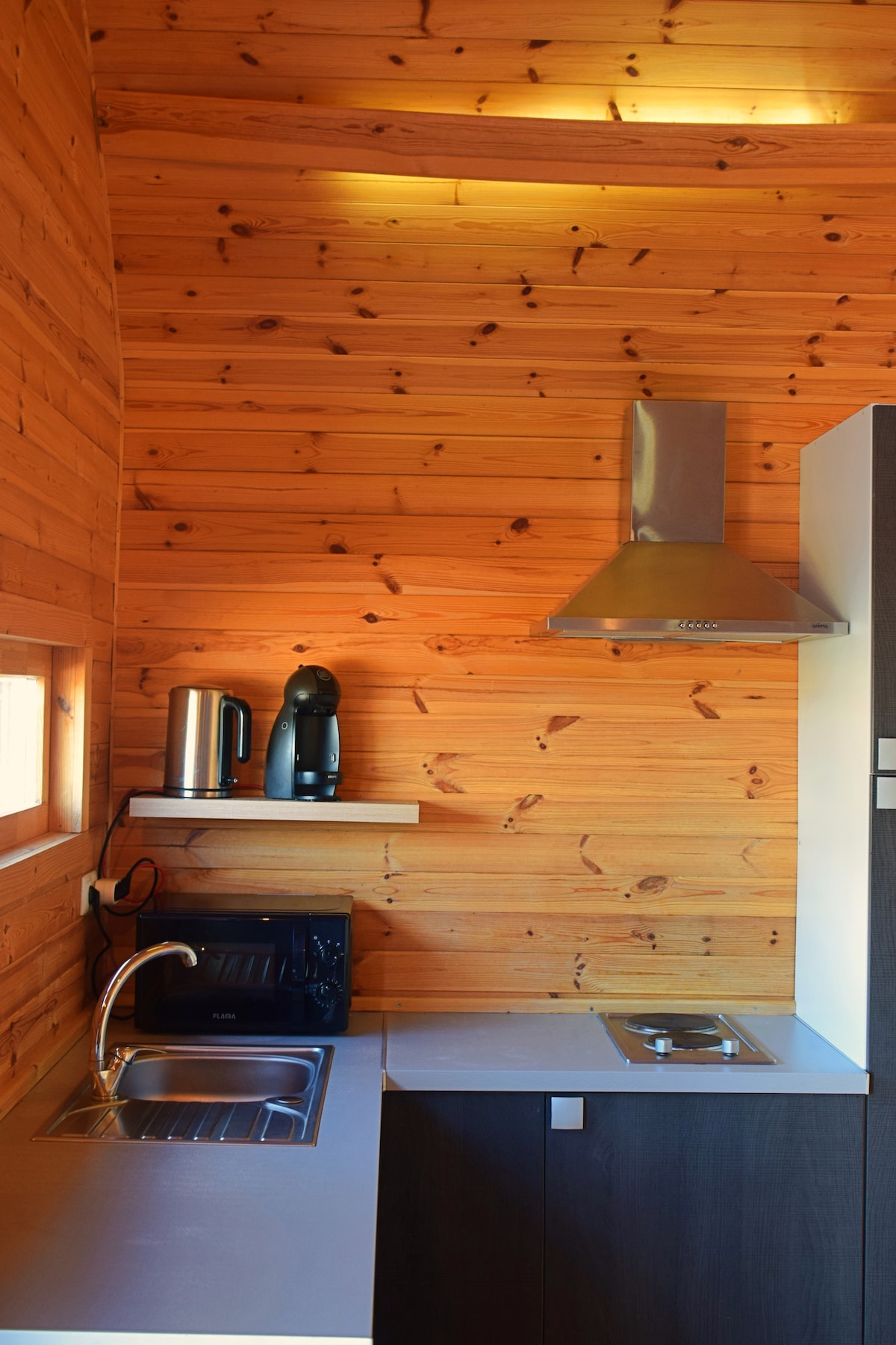 A compact kitchen area is presented, featuring wooden walls that add warmth. Modern appliances include a kettle, coffee maker, and a microwave, along with a stainless steel sink and a stovetop. Ample counter space supports meal preparation.