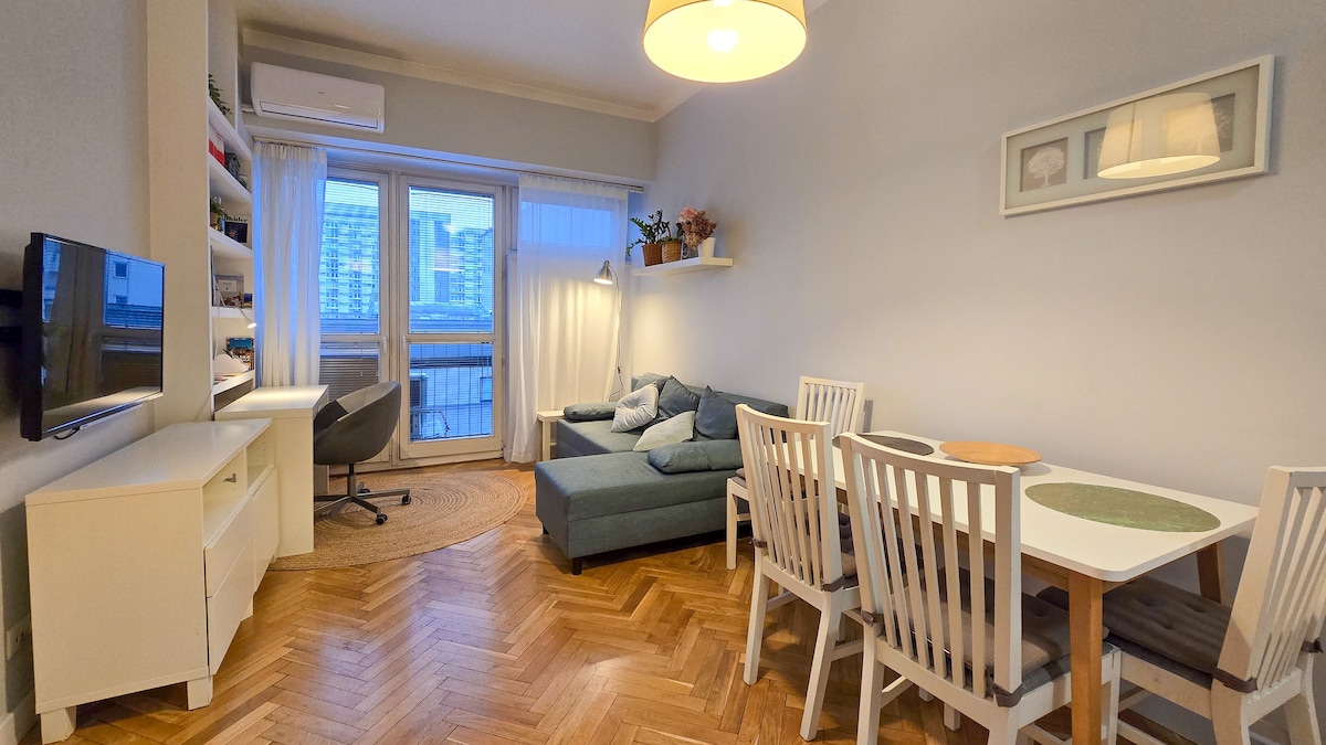 A bright living space displays a comfortable sectional sofa beside large windows, allowing natural light to fill the room. A dining table with white chairs is positioned near a simple, modern desk. Wooden flooring adds warmth to the inviting atmosphere.