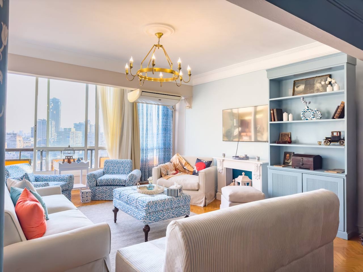 The living area features a spacious arrangement of light-colored sofas and patterned armchairs, offering comfortable seating. Large windows provide abundant natural light and views of the city. A chandelier hangs from the ceiling, while decorative shelves display various items, adding a touch of elegance to the space.