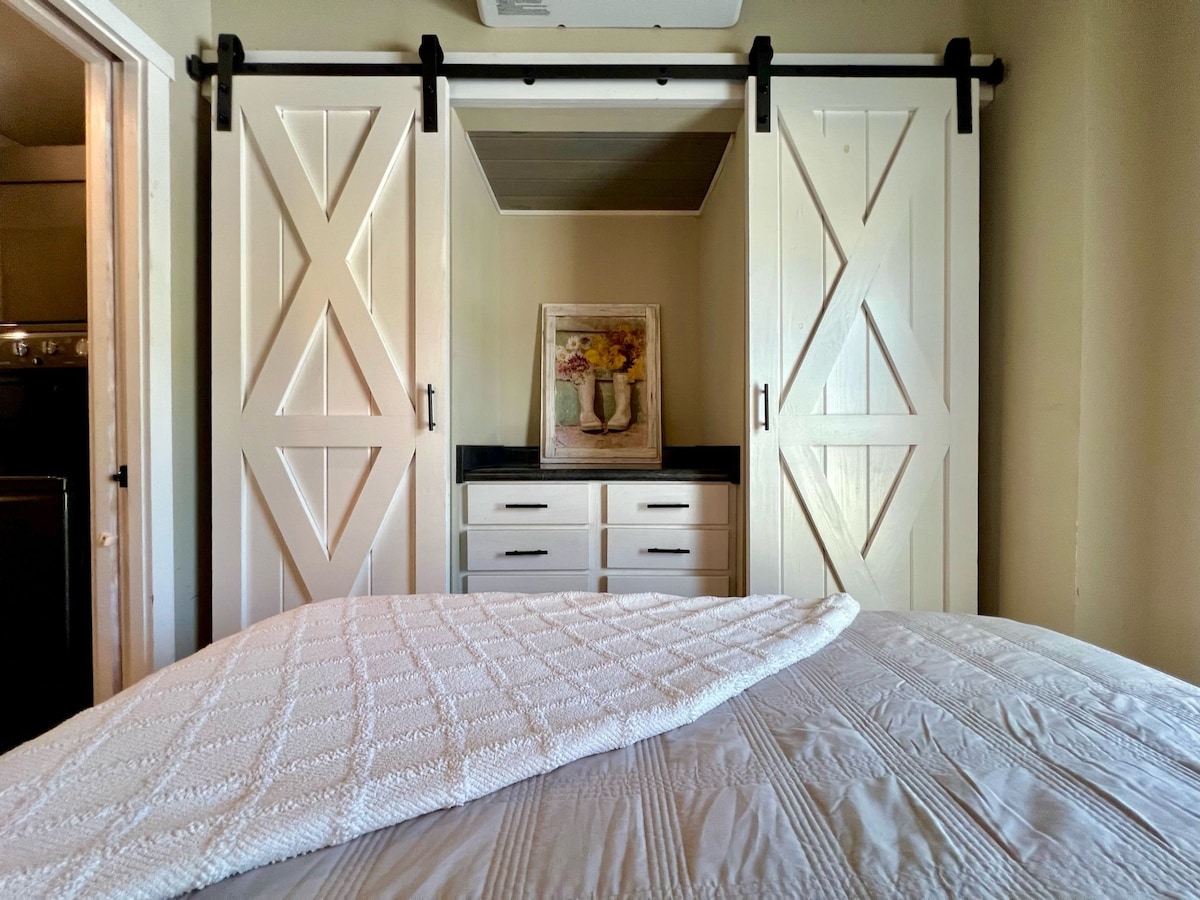 A cozy bedroom space is featured, showcasing a spacious bed covered with a textured white blanket. Sliding barn doors are positioned in the background, revealing storage options. A floral artwork is displayed above a dresser, adding a touch of elegance to the room.