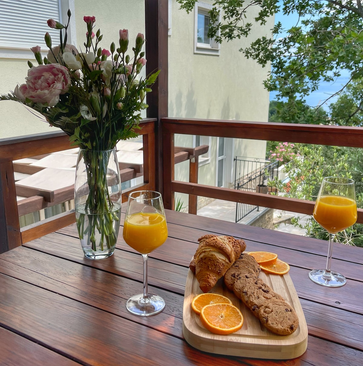 A sunny terrace features a wooden table set with two glasses of orange juice and a wooden platter of croissants and orange slices. A vase of fresh flowers adds a touch of nature, while the backdrop shows greenery and a glimpse of the sea.