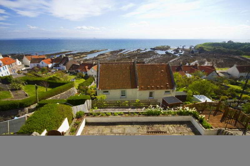 A panoramic view captures the coastal landscape, featuring charming houses with red roofs, rocky shorelines, and the shimmering sea. Green terraced gardens lead down to the shoreline, enhancing the outdoor space. The sky is bright with scattered clouds, adding to the serene atmosphere.