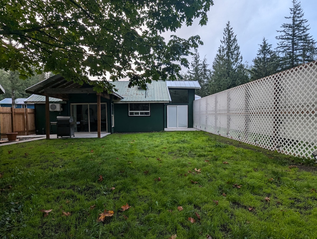 A spacious yard is depicted, featuring a grassy area surrounded by trees. A covered patio is visible, along with a green exterior of the guest house. A white lattice fence provides separation and is seen to one side of the yard, enhancing the outdoor space.