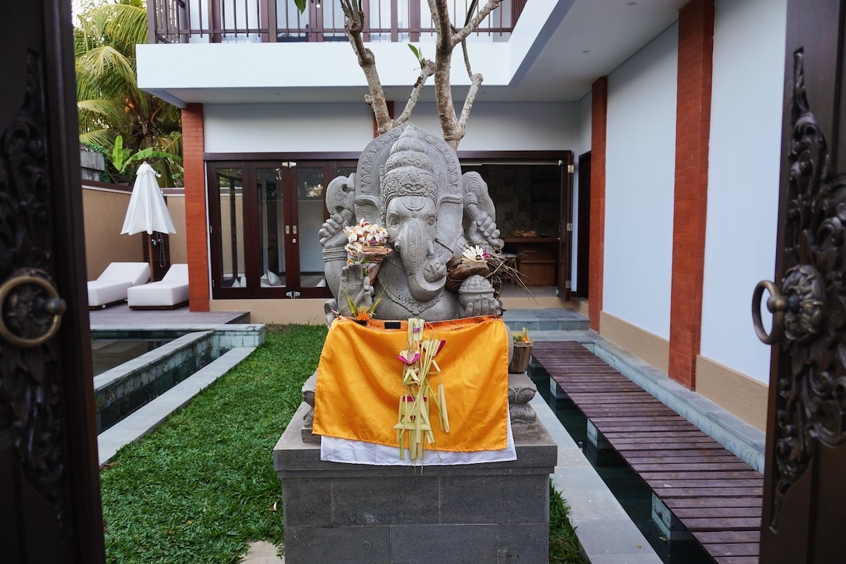 A majestic Ganesha statue is prominently displayed at the entry, adorned with offerings. The surrounding area features lush green grass and a reflective pool, creating a serene environment. The structure of the house is visible in the background, with shaded seating areas enhancing the tranquil feel.