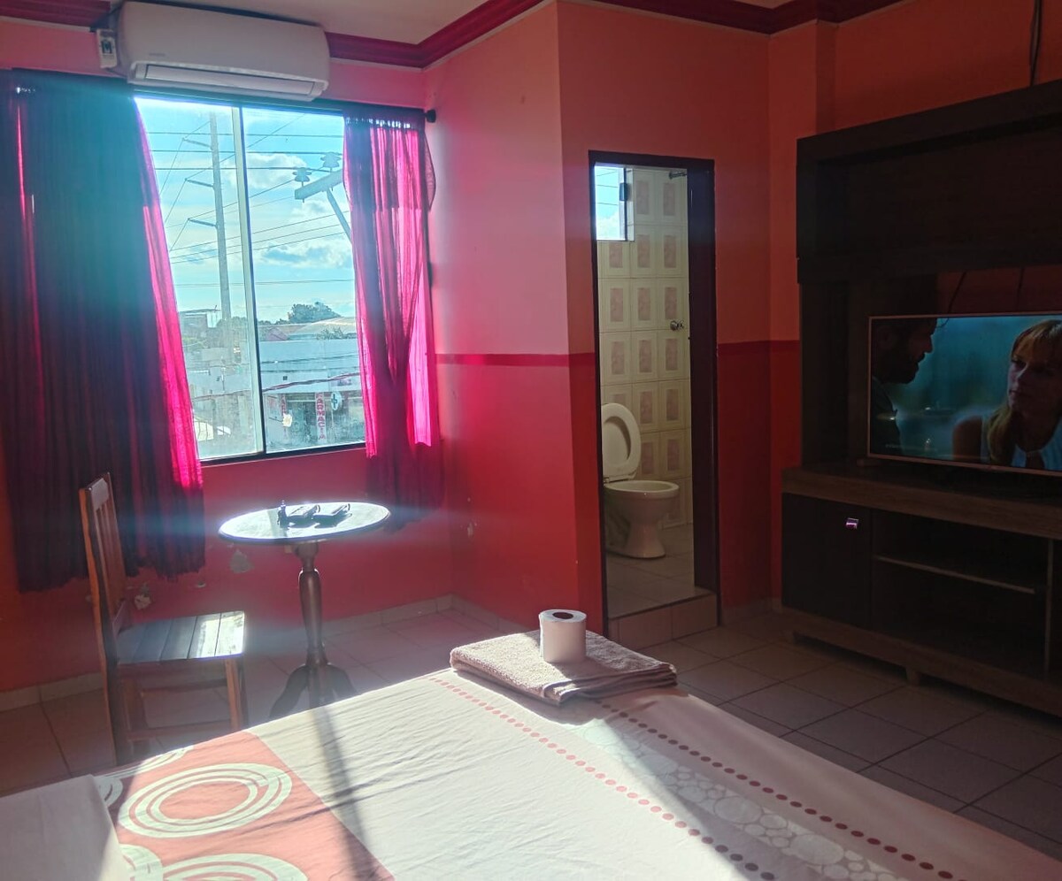 A bright room features a spacious bed with crisp linens, complemented by a television mounted on a dark wood unit. A small circular table and chair are positioned near the window, which is adorned with flowing red curtains, allowing natural light to fill the space.