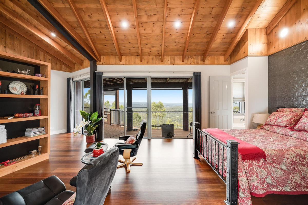 A spacious room features vaulted wooden ceilings and large windows that provide expansive views. A neatly made bed with a patterned red coverlet is complemented by a stylish chair and a bookshelf, creating a comfortable and inviting atmosphere.